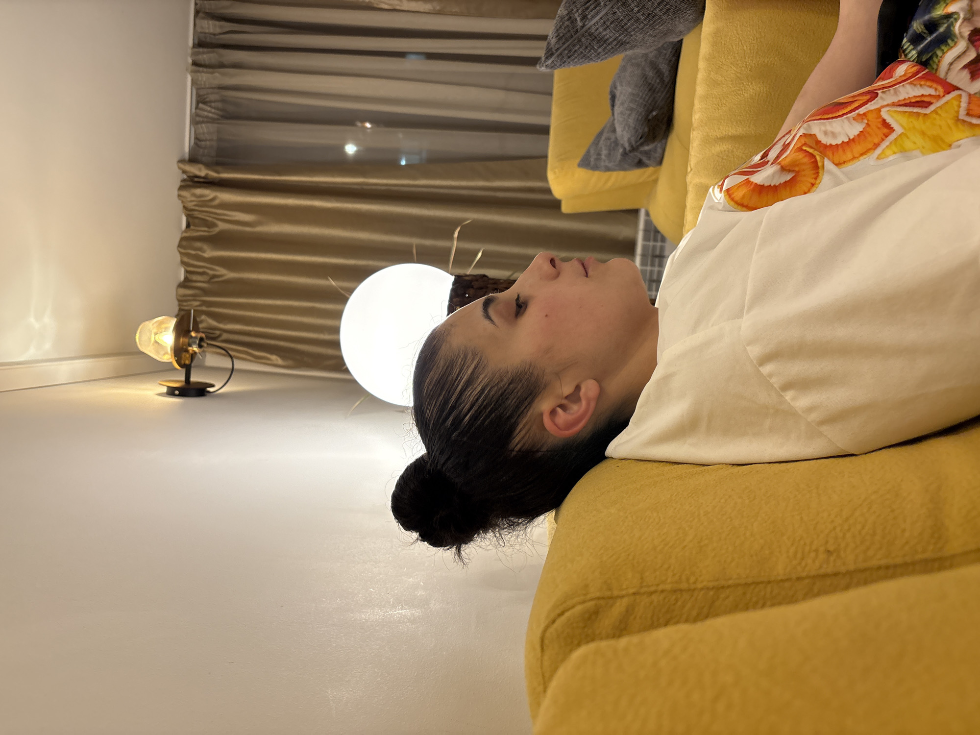 A woman is lying on a yellow sofa. The sofa is positioned in a room with a white floor. Near the sofa, there are two lamps, one with a spherical shade and another of a smaller, more decorative design. In the background, there is a window covered with golden-toned curtains. Based on the architectural style and the general features, it is difficult to definitively ascertain if this is Sofia, Bulgaria without more specific visual or contextual cues.