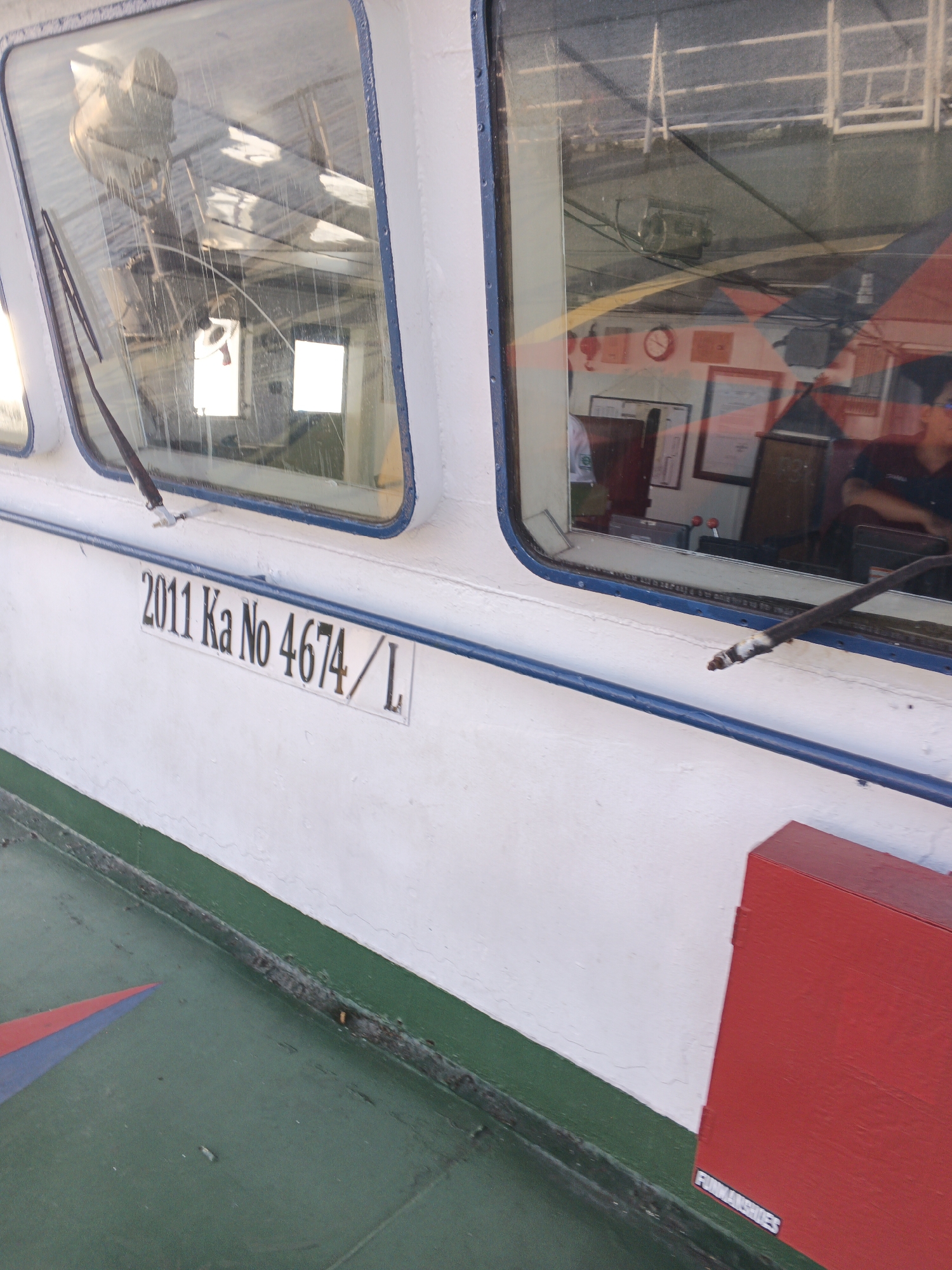 A close-up, eye-level shot captures a white boat hull with the registration number "2011 Ka No 4674 / L" painted in black on a white rectangular plate. Above the registration, a large, angled window reveals the interior of the boat's cabin. Reflections on the window obscure many details, but a large mechanical object, possibly a radar or antenna, is visible in the upper left. The cabin interior shows various instruments, a clock, and framed items on the walls. In the right side of the cabin, a person wearing a dark shirt is visible, appearing to be seated and looking away from the camera. The boat's deck, painted in a dark green, is visible in the lower portion of the image, with a red and blue triangular design. A red metal box, possibly a storage unit or fire extinguisher box, is situated on the deck near the hull. The boat is likely docked or anchored in the waters of Melaya, Indonesia, a coastal district known for its fishing communities and marine activities.