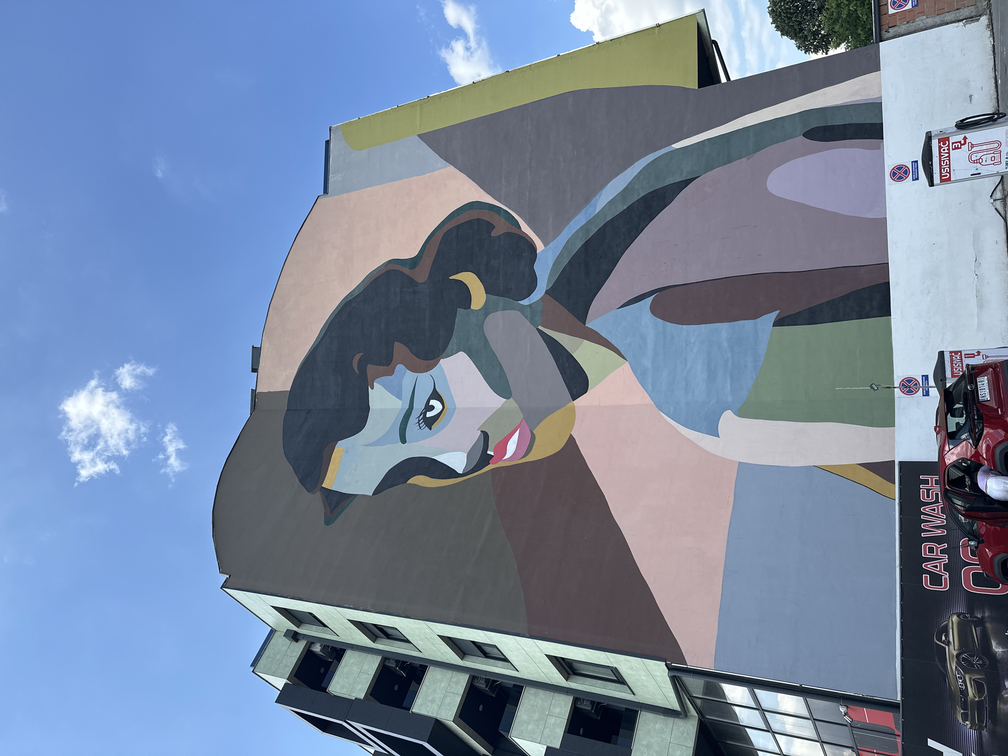 A street-level, upward-angled view captures a multi-story building in Novi Sad, Serbia, adorned with a mural of a woman's face in a stylized, geometric art form. The building's lower stories are modern, featuring black and grey cladding and numerous windows, while the mural covers a curved section of the upper stories, employing a palette of beige, brown, green, blue, and black.
To the right of the building, there is a vertical sign that reads "CAR WASH." A red car is positioned partially in view behind the sign. The sky above is a bright blue with scattered white clouds. The USISSIVAC car wash units are also on the right.