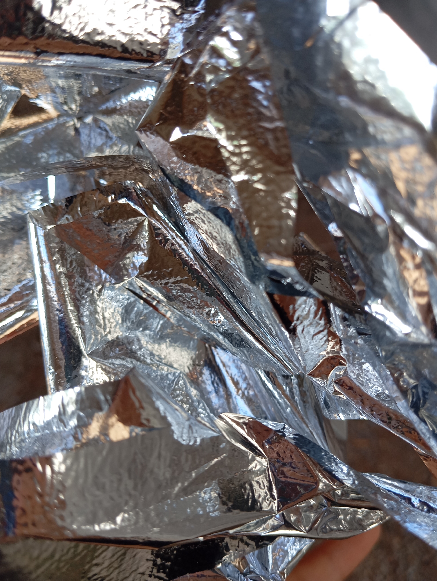 This image is a close-up, abstract shot of crumpled, shiny, silver material, likely a mylar emergency blanket. The material is highly reflective, capturing light and creating bright highlights and deep shadows that obscure any distinct shapes or objects. The texture of the material is emphasized, with numerous folds, creases, and wrinkles visible. The lighting appears to be natural, possibly sunlight, judging by the bright reflections and the warm tones peeking through some of the creases. The background is out of focus, showing hints of a brown, earthy surface and possibly some greenery, suggesting an outdoor setting. No people are clearly visible, although a hint of a hand or finger can be seen in the lower right corner, indicating someone is holding or manipulating the material. No text is present. The overall impression is one of texture and light play on a reflective surface, with an abstract and somewhat disorienting composition due to the close-up perspective and the material's crinkled nature.