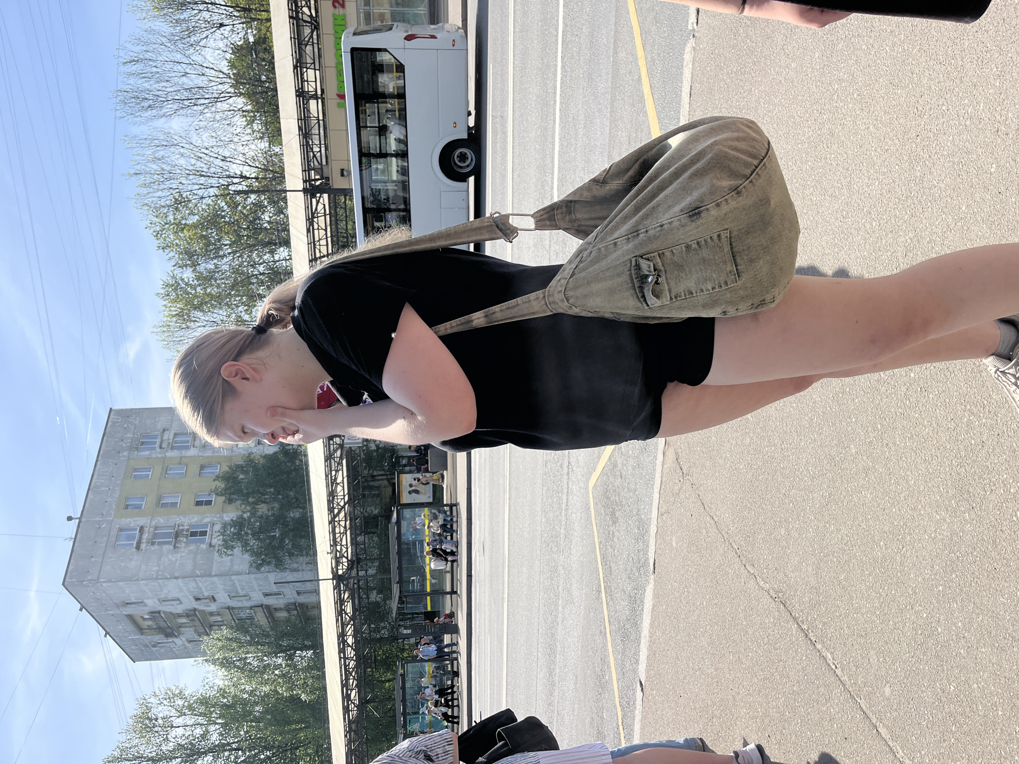 A person is standing on a sidewalk next to a bus lane in Saint Petersburg, Russia. The person is wearing a black t-shirt, black shorts, and carrying a light green shoulder bag. The person is fair-skinned with light blonde hair pulled back in a ponytail. In the background, there is a bus, a bus stop with people waiting, and a large, multi-story building with yellow accents. Trees line the street. The sidewalk is concrete and appears to be cracked in places. A second person is partially visible in the bottom of the image.