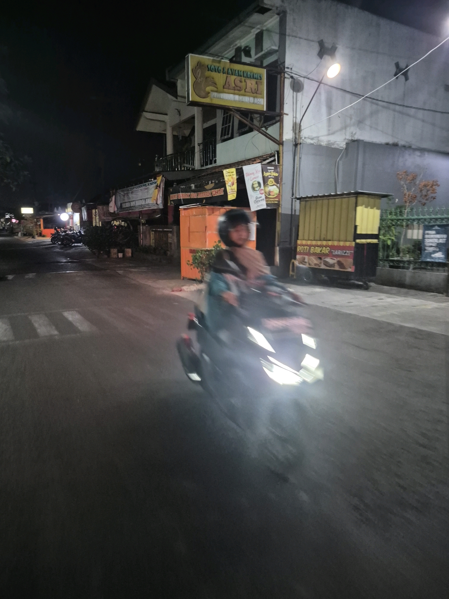 In Pabelan, Indonesia, at night, a person rides a motorcycle on a street. The motorcycle's headlight is illuminated, casting a bright glow. The person is wearing a helmet and is blurred due to motion. To the right of the street, a food stall with a yellow and black striped facade is visible, advertising "ROTI BAKAR TARIZZE." The stall has images of bread and food items. Behind the stall, a green fence and some trees can be seen. On the left side of the street, a building with a yellow sign reading "SOTO & AYAM KREME ASIH" is present. The sign is illuminated, and it appears to be a food establishment. There are other businesses and parked motorcycles further down the street, suggesting a commercial area. The street is paved, and a pedestrian crossing is marked with white stripes. The overall environment appears to be a moderately lit urban street scene at night.