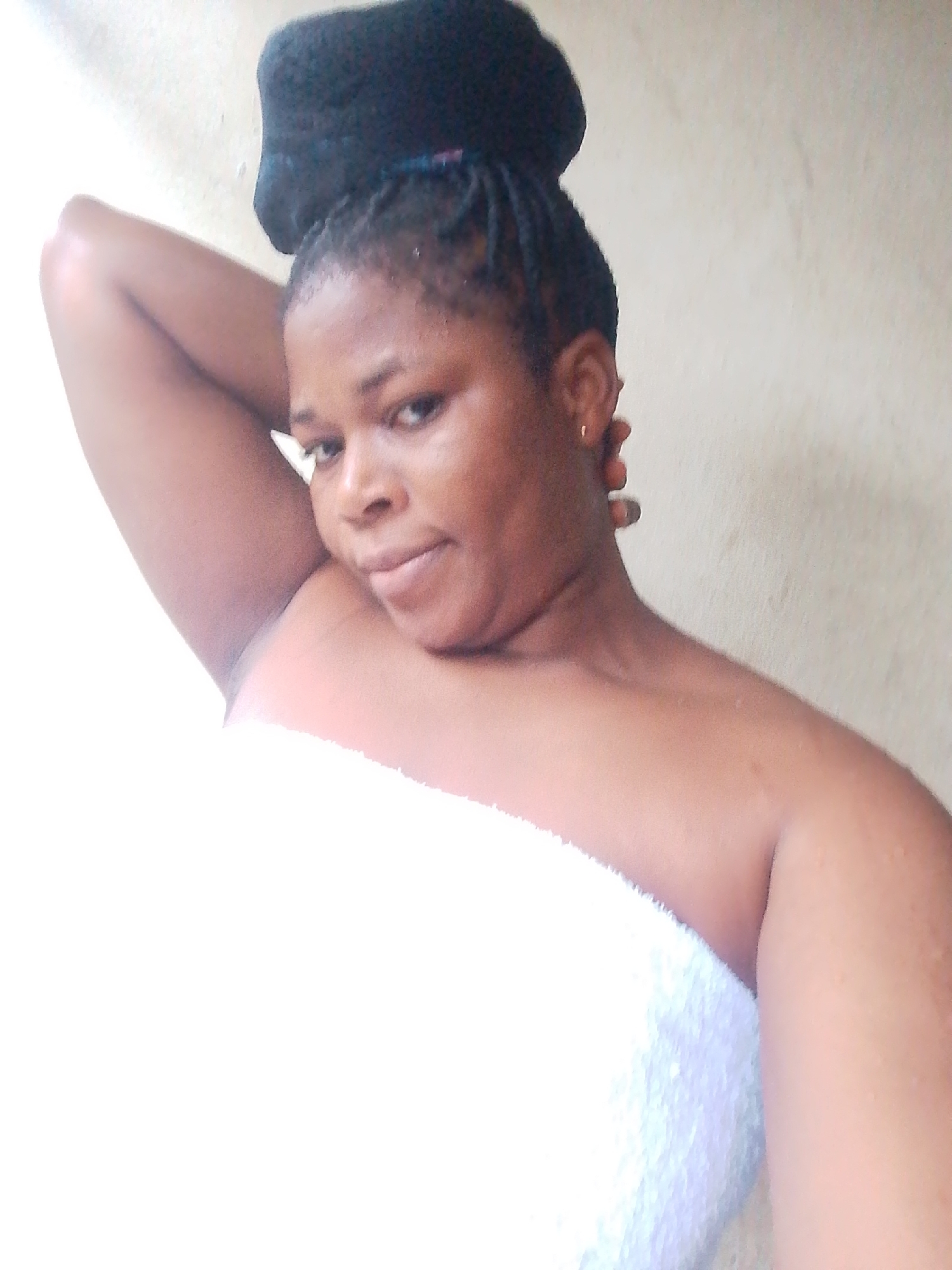 A medium-shot photo, taken from a slightly high angle, shows a Black woman from the chest up. She is wearing a white towel around her chest and has her left arm bent behind her head. Her hair is pulled up into a bun on top of her head, and she has a small gold earring in her left ear. She is looking at the camera with a relaxed expression. The background is a plain, light-colored wall. The lighting is bright, with the left side of the image appearing overexposed.