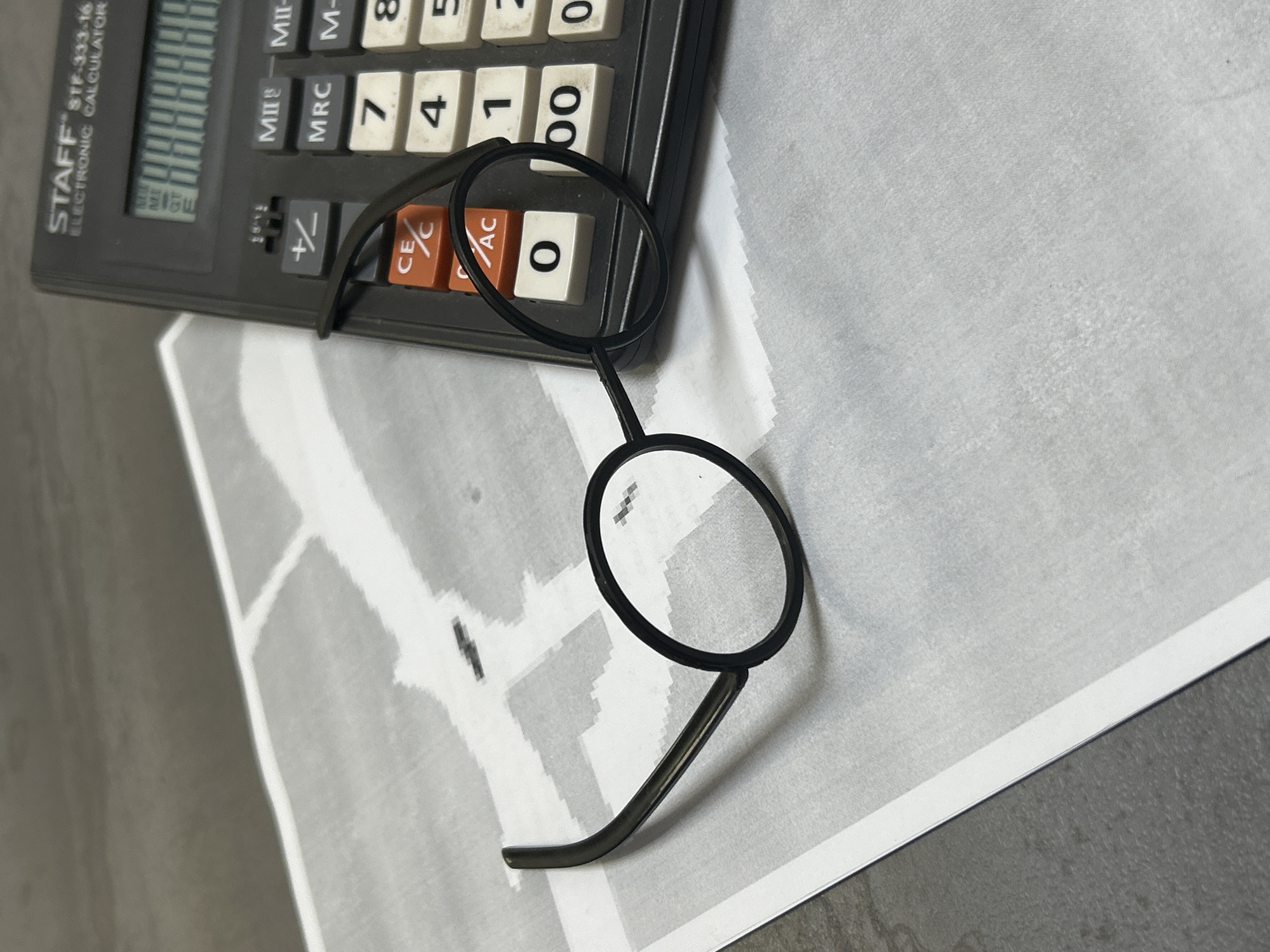 The image presents a flat lay composition of a dark gray electronic calculator, a pair of black-framed round glasses, and a piece of paper, all resting on a plain gray surface. The scene appears to be an indoor setting in Chaikovsky, Russia, though no specific geographical markers are visible.
The calculator, branded "STAFF STF-333-16 ELECTRONIC CALCULATOR," occupies the upper left portion of the frame. Its dark body features white number keys (0, 00, 1, 4, 5, 7, 8) and orange function keys ("CE/C", "AC"). Other visible labels on the keys or above them include "MII-", "MRC", "M-", "NEW", "NLE", "GT", and "MILE". The calculator's display shows faint, segmented characters that are not clearly legible.
Overlapping the calculator and extending onto the paper are the round glasses. They have thin, black frames and are designed with a single, simple bridge connecting the two circular lenses. One arm of the glasses is visible, curving away from the lenses.
Beneath the calculator and glasses is a sheet of paper featuring an abstract pattern of light gray and white blocks or shapes, creating a somewhat geometric or tiled appearance. The entire arrangement is situated on a smooth, uniform gray surface, possibly a desk or floor, which provides a neutral background for the objects. No specific activity is depicted; the objects are simply arranged.