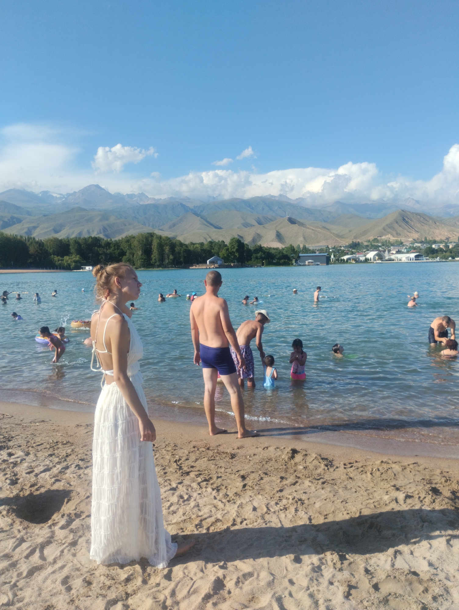 The image shows a sunny day at a beach in Cholpon-Ata, Kyrgyzstan, with people swimming and relaxing in the clear blue water of Lake Issyk-Kul. The background features lush green trees and a distant mountain range under a bright blue sky with scattered clouds. In the foreground, a woman in a long white dress stands on the sandy beach, facing away from the camera. In the mid-ground, several people are visible in the water, including adults and children. Some are swimming, while others are standing or sitting near the shore. A man in blue swim trunks is standing in the water near the woman in white. The beach is made of light brown sand with some footprints and shadows. The water is calm with gentle waves lapping the shore. The overall scene depicts a lively summer day at a popular tourist destination.