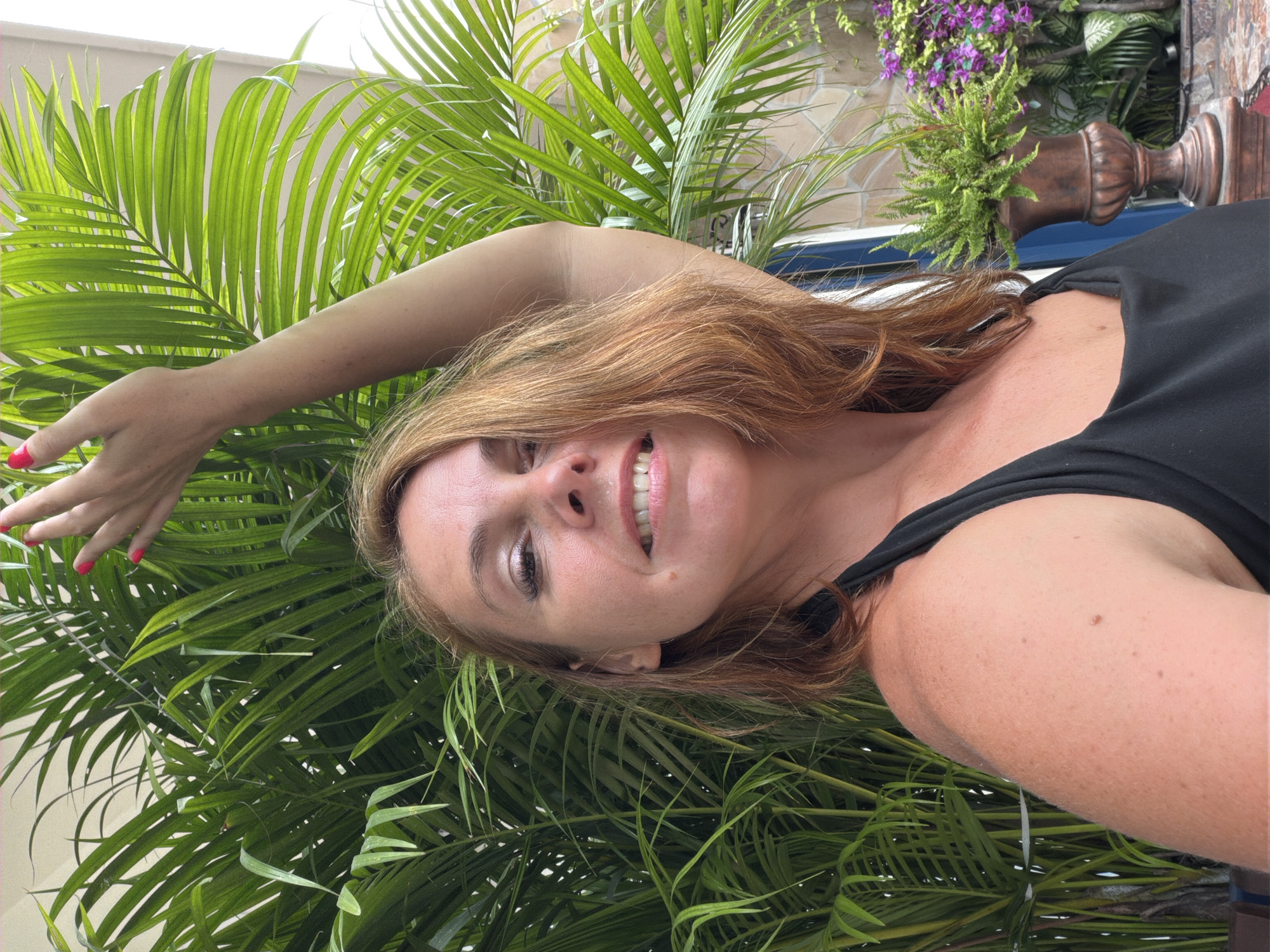 A woman with auburn hair and fair skin, wearing a black halter top, is pictured outdoors with a large green palm plant behind her. She is looking at the camera with a wide smile, her right arm raised. Her fingernails are painted red. To the right of the palm plant, a potted fern and purple flowers are visible. The background suggests an outdoor patio or garden setting.