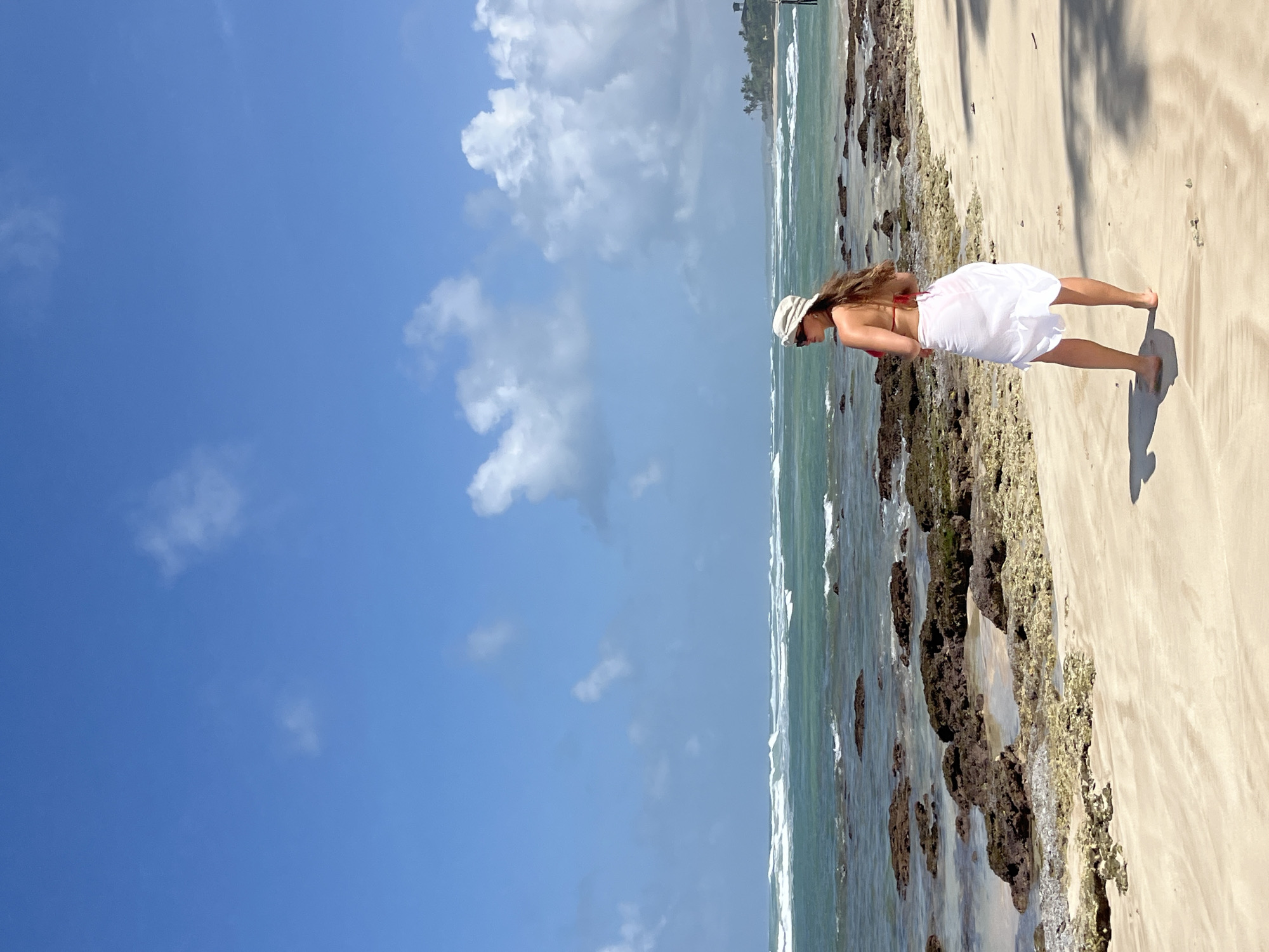 A woman stands on a sandy beach facing the ocean in Kathaluwa, Sri Lanka. She is wearing a red bikini, a white sarong, and a light-colored hat. Her back is to the camera as she looks out at the turquoise water, which has white waves breaking on a rocky shoreline. The sky is a bright blue with scattered white clouds. The sand is light brown and there are shadows cast on it by palm trees that are not fully visible. The overall scene is one of a tropical beach vacation.
