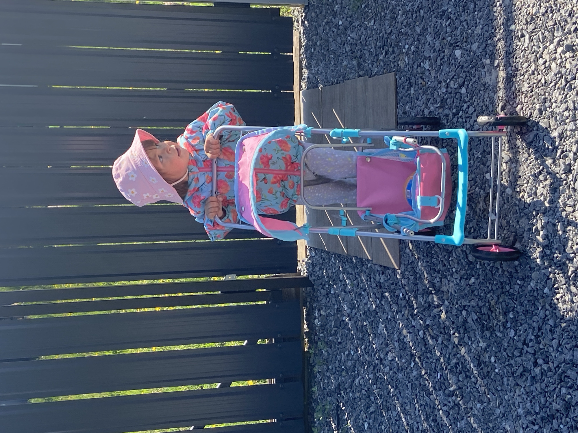 A young child wearing a pink bucket hat and a floral-patterned jacket sits in a toy stroller with a pink and blue color scheme. The stroller is positioned on a gravel surface adjacent to a dark-colored fence. The scene is outdoors, illuminated by sunlight that creates shadows on the gravel. The child is looking to the left. While the image's metadata is unavailable, the query specified the city of Tyumen, Russia as the location. Therefore, the scene is presumed to be in Tyumen, Russia.