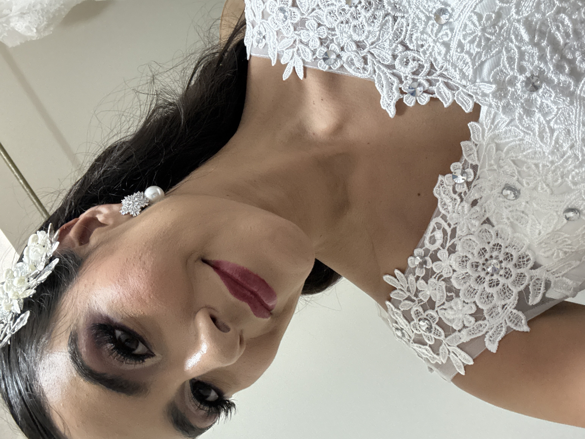 A close-up, slightly angled portrait shows a woman wearing a white wedding dress with intricate lace detailing and small sparkling embellishments around the neckline. She has dark hair and wears a silver headpiece with small white pearls, along with matching pearl and diamond-like earrings. Her makeup is professionally done, featuring dark eyeshadow, defined eyelashes, and deep red lipstick.
While the image provides details about the woman and her attire, it does not contain any information that indicates the location as Puebla, Mexico. The background is a plain white wall, offering no visual clues about the city or its environment.