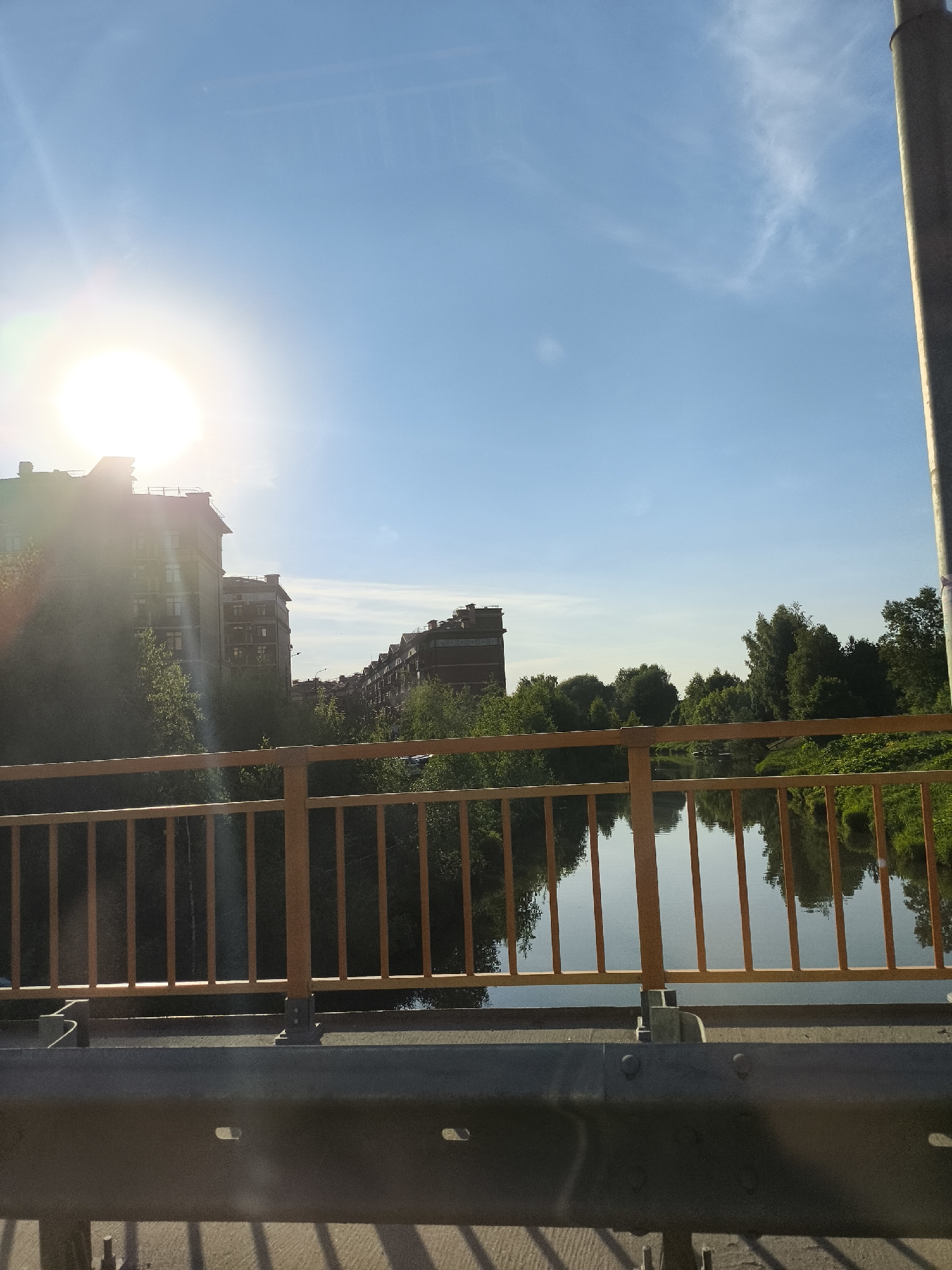 The image displays an urban landscape scene captured from a bridge in the city of Unknown, Unknown. The scene is illuminated by bright daylight, originating from an overexposed sun situated in the upper-left quadrant, which produces significant lens flare. The sky is a clear, light blue with minimal, wispy cloud formations.
In the mid-ground, a calm body of water, identified as a river, flows horizontally across the frame, reflecting the blue sky and the surrounding green vegetation. On the left bank of the river, several multi-story residential buildings with dark roofs are partially visible through a dense canopy of green deciduous trees. These structures appear to be modern apartment complexes. The far bank of the river is heavily forested with lush green trees. A dark grey, vertical utility pole or lamppost is visible along the extreme right edge of the frame.
The foreground is dominated by elements of the bridge structure. An orange-yellow metal balustrade, composed of vertical pickets and horizontal rails, extends horizontally across the mid-foreground. Below this balustrade, a dark grey metal crash barrier or guardrail, secured with visible bolts and featuring elongated rectangular apertures, spans the lower portion of the frame. The paved surface of the bridge deck is visible beneath the balustrade and guardrail, exhibiting distinct grid-like shadows in the bottom-most section of the image. No individuals or active movement are present within the frame.