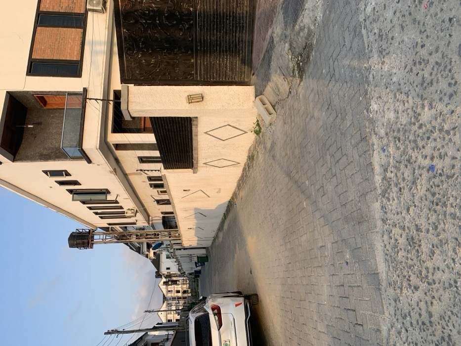 A building is visible on the left side of the shot. It has multiple windows and a dark metal gate. The facade is decorated with diamond-shaped patterns. Next to the building is a paved street with a white car parked on the side. Further down the street, more buildings can be seen. A water tank stands tall on a metal structure, and overhead power lines stretch across the sky. The sky is light blue.