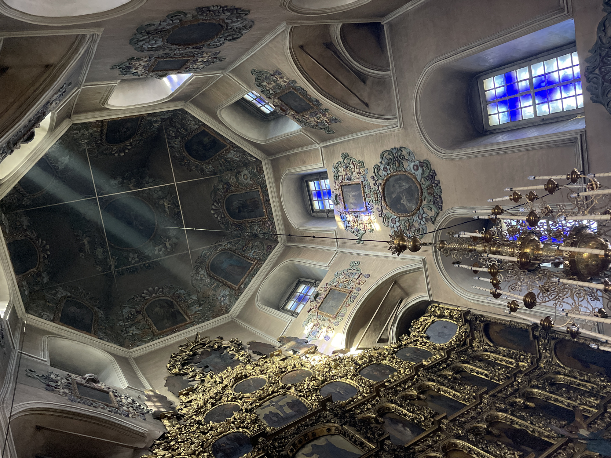 The image displays the ornate interior of a grand building, likely a church or cathedral, viewed from a low angle looking towards the ceiling. The central feature is a multi-faceted, possibly octagonal, domed ceiling adorned with painted panels depicting figures or scenes, set within intricate dark blue or green decorative borders. Rays of light, possibly sunlight, stream through the space, illuminating the air.
Several arched windows are visible along the walls, which are painted in a light brown or beige hue. One prominent window in the upper right features distinct blue stained glass arranged in a cross-like pattern. A large, elaborate multi-tiered golden chandelier with numerous light fixtures hangs from the ceiling.
In the lower right foreground, a magnificent, heavily gilded structure dominates, appearing to be an iconostasis or a richly decorated altar screen. It is adorned with intricate golden carvings and features numerous smaller framed paintings or icons. Throughout the interior, decorative elements in gilded or painted stucco, frames, and ornate motifs in muted blue, green, and gold tones enhance the opulent atmosphere.
No people or specific activities are depicted. The presence of light rays suggests it is daytime. No visible text is present. The architectural style, with its extensive ornamentation and gilding, evokes a Baroque or Rococo aesthetic.