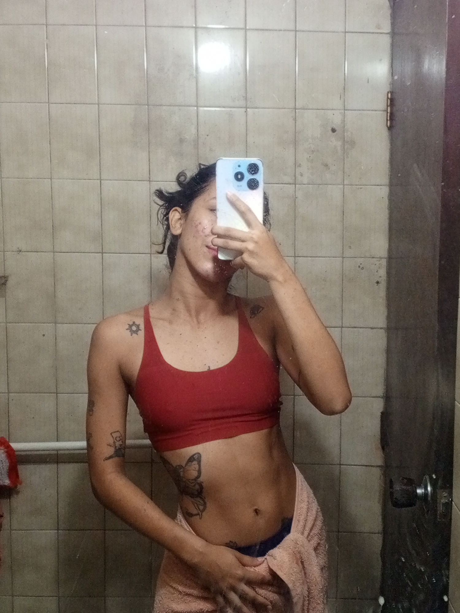 A young adult stands in a bathroom, holding a smartphone in front of their face to take a selfie. The person is wearing a red sports bra and a light pink towel wrapped around their waist. Tattoos are visible on the person's shoulders, arms, and torso, including a butterfly tattoo on their side. The bathroom features light-colored square tiles on the walls. A towel rack with a red and white towel on it is visible to the left. The bathroom door, which is dark and worn, is visible to the right. The location is possibly a private residence in Porlamar, Venezuela, although this is not definitively confirmed by visual elements in the image.