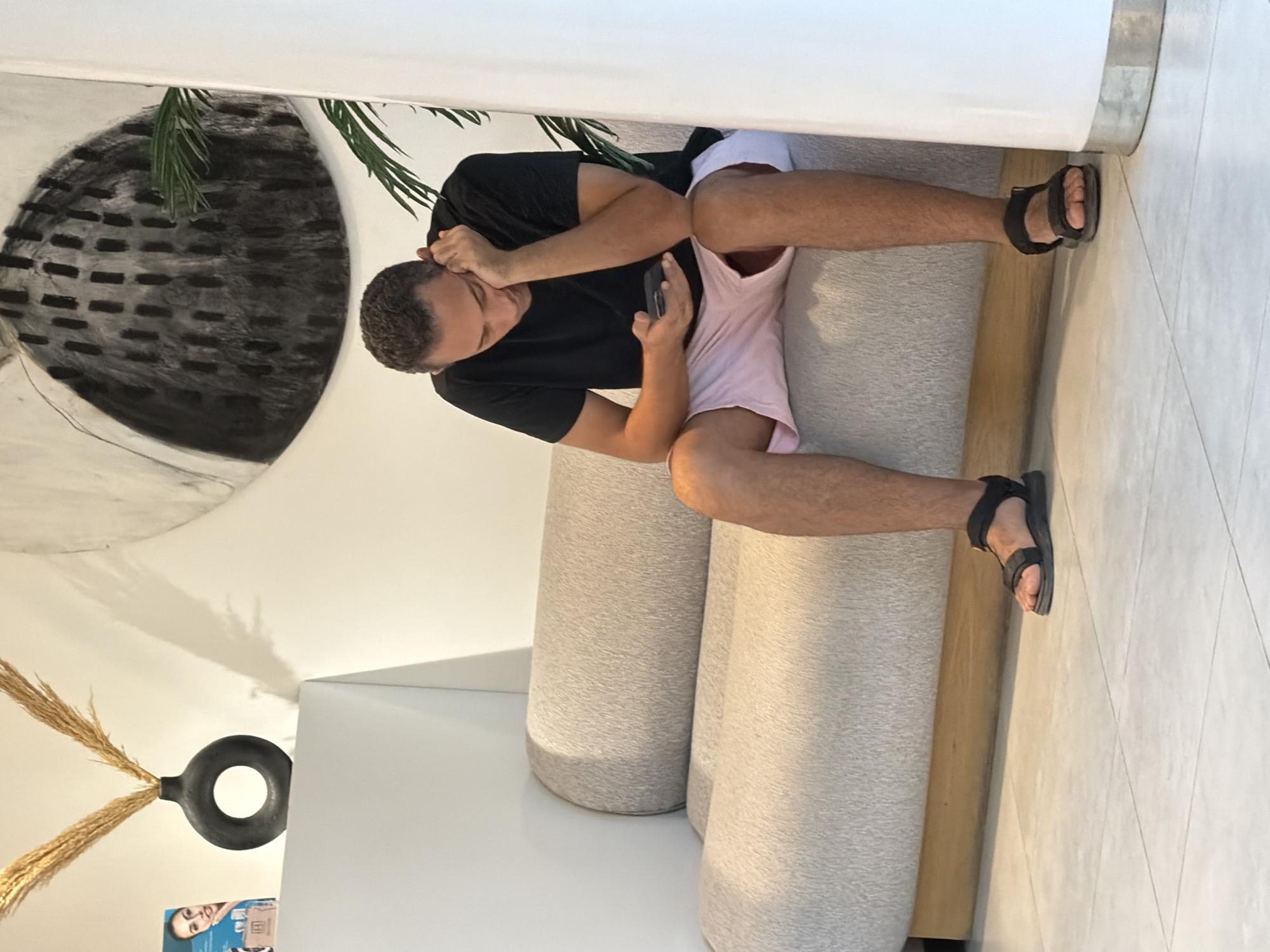A man with short, curly hair wearing a black t-shirt and light pink shorts is sitting on a light-colored, curved sofa in a room that appears to be a waiting area or lounge. He is holding a smartphone in his right hand and resting his head on his left hand, with his gaze directed downwards at the phone. The man is wearing black sandals.
In the background, there is a large, abstract charcoal drawing on the wall, predominantly black with white and gray accents, depicting a circular, textured shape. To the left of the drawing, a dark gray, circular vase with a hole in the center contains dried wheat-like stalks. Next to the vase is a white shelf with a book or magazine featuring a woman's face on its cover. A large, white cylindrical object is visible on the right side of the frame. The flooring is tiled in a light neutral color.
The image does not contain any specific visual cues to determine its location in Sharm El Sheikh, Egypt.