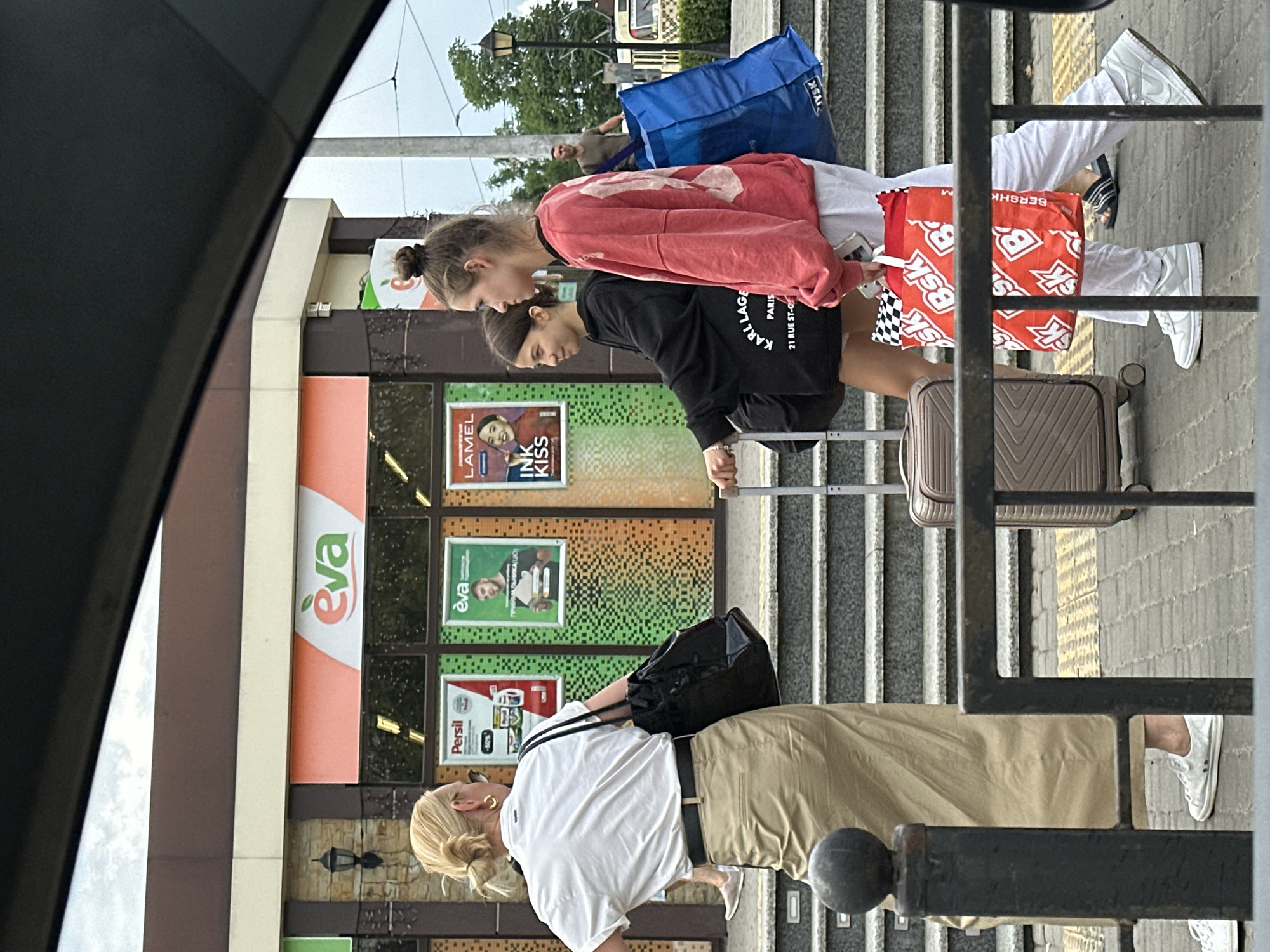 The image depicts a street scene in Odesa, Ukraine, near a "EVA" store. In the foreground, a woman with blonde hair pulled back in a bun and wearing a white shirt and a tan skirt is walking away from the camera, carrying a black bag over her left shoulder. To her right, two younger women are walking towards the camera. The woman on the left wears a black hoodie and the woman on the right wears a red sweatshirt. Both are carrying luggage. The woman on the right is carrying a blue shopping bag and a red shopping bag with "BSK" written on it. Behind them, a man in a dark shirt is walking in the background. The scene is set on a paved walkway with steps leading up to the store. The weather appears overcast.