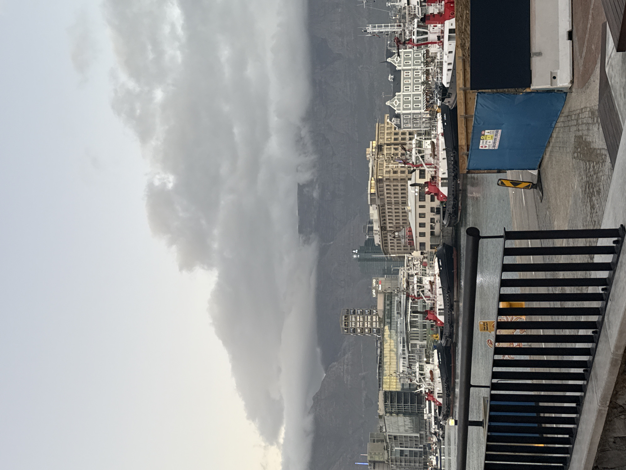 The image depicts Cape Town's harbor, obscured by low-hanging clouds enveloping Table Mountain in the background. Tall buildings line the waterfront, with cranes visible among the structures, indicating ongoing construction or port activity. A section of black metal railing is in the foreground, and a blue tarp-covered structure is visible along the waterfront.