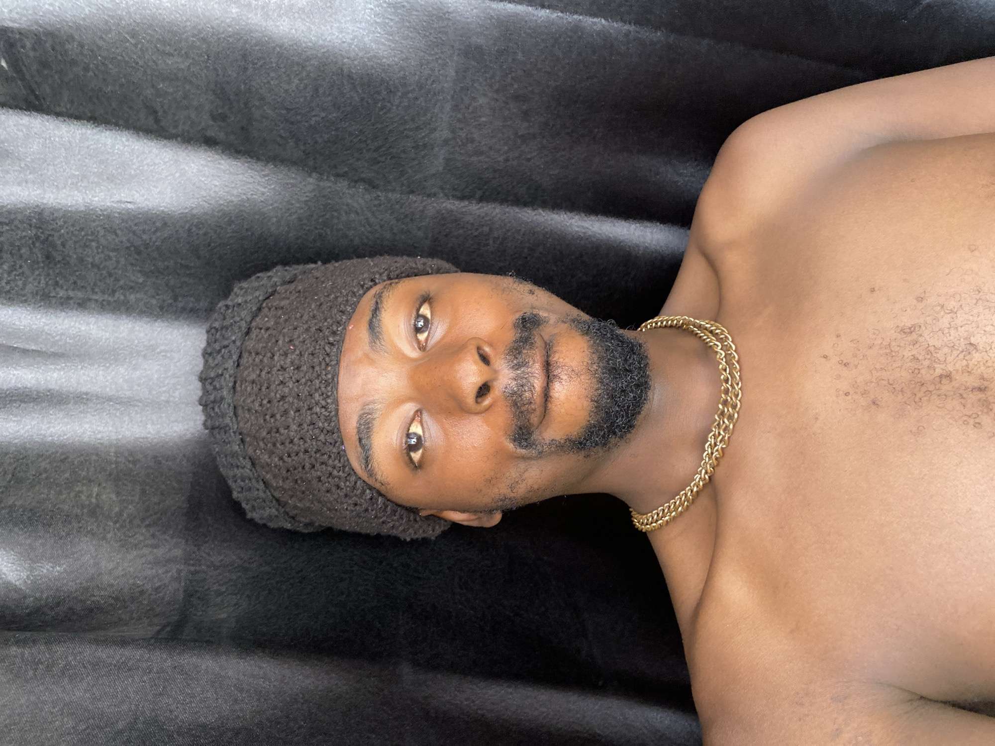 A head-and-shoulders portrait depicts a dark-skinned man lying down against a dark, textured fabric background. He is looking directly at the camera with a neutral expression. The man wears a dark, possibly black or charcoal gray, knitted hat or beanie. A prominent gold-colored chain necklace with multiple links is visible around his neck, resting on his bare chest. He has a short, well-groomed beard and mustache. The background fabric shows soft folds and varying shades of dark gray and black, suggesting a bed, couch, or draped material. The lighting appears soft, highlighting the subject's features and the texture of the fabric. No specific location cues for Barakin Akawo, Nigeria, are discernible from the visual information alone. There is no visible text.