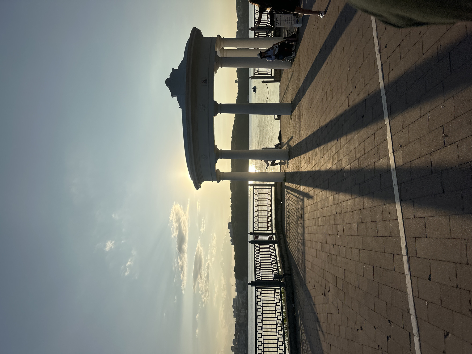 The image depicts a scenic view of the Ternopil Pond in Ternopil, Ukraine, during sunset. A white rotunda with classical columns stands prominently in the foreground, casting long shadows across the paved walkway. The pond stretches out beyond, with a treeline and city skyline visible on the opposite bank. Several people are present in the scene: a musician with a hat and guitar sits on a chair in front of the rotunda, a woman in a black dress stands on the right, and another person is visible in the distance. The sun, low in the sky, creates a warm, golden light and a striking silhouette effect. The atmosphere is serene and picturesque, typical of a late summer or early autumn evening.