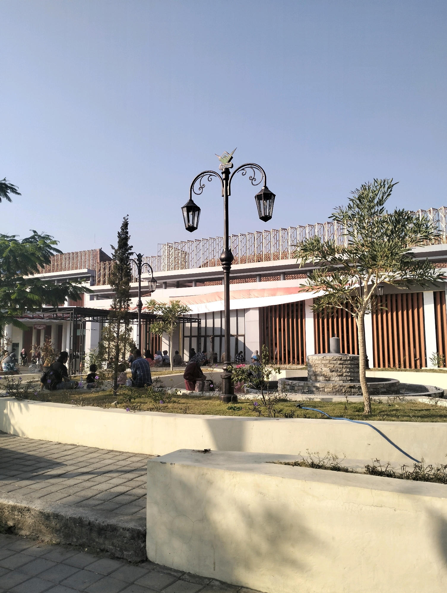 The image depicts an outdoor area with a light post in the foreground, featuring two lamps and a decorative bird figurine at the top. The background shows a modern building with vertical wooden slats and a second-story walkway. Several people are scattered throughout the scene, some sitting on the grass and a low wall, others appearing to converse or relax. A small tree with thin branches and green leaves stands near a stone fountain in the mid-ground. The ground in the foreground is paved with interlocking rectangular stones. The sky is clear and blue, suggesting daytime. The location is identified as Bangunsari, Indonesia.