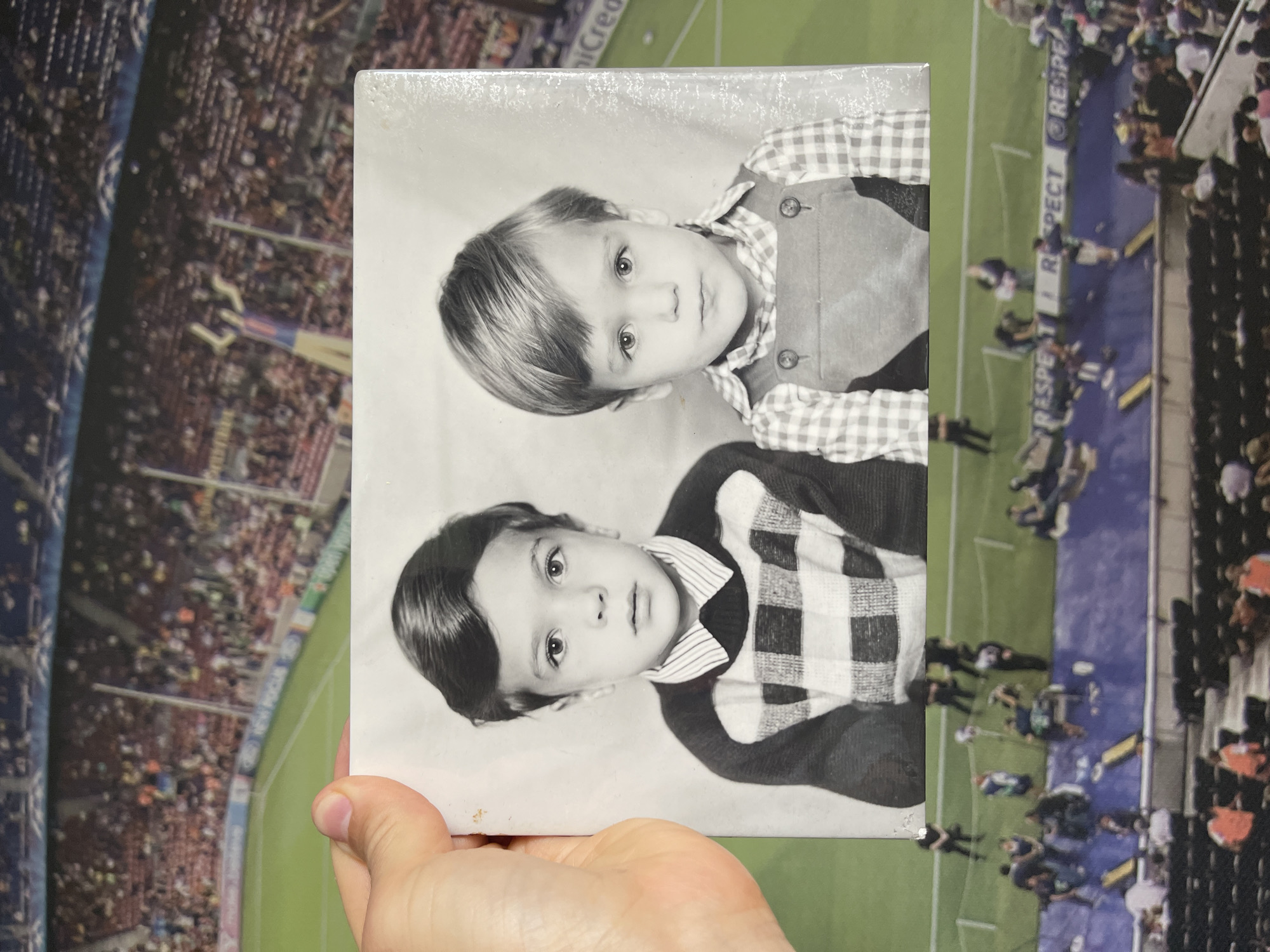 A hand holds an old black and white photograph of two young children, possibly brothers, against a vibrant, out-of-focus background of a sports stadium. The photograph and the background are both oriented vertically, suggesting the original capture was rotated 90 degrees clockwise.
In the black and white photograph, two boys, appearing to be toddlers or preschoolers, are depicted. The upper child has lighter hair, neatly parted, and wears a gingham-checked shirt under a buttoned vest. The lower child has darker, slicked-back hair with a distinct parting, and wears a striped collared shirt beneath a checkered sweater. Both children look directly at the camera with neutral expressions. The photograph shows some minor wear and dust.
The background is a colorful, well-lit sports stadium, likely a football (soccer) venue. A green pitch is visible, surrounded by a blue track or perimeter area. The stands are packed with numerous spectators, indicating a live event. Advertising hoardings around the pitch clearly display the word "RESPECT" multiple times, along with a partial logo that includes "iCred". The stadium appears to be an outdoor setting, potentially under evening lights or bright daytime conditions. The hand holding the photograph is light-skinned, visible at the bottom right of the print.