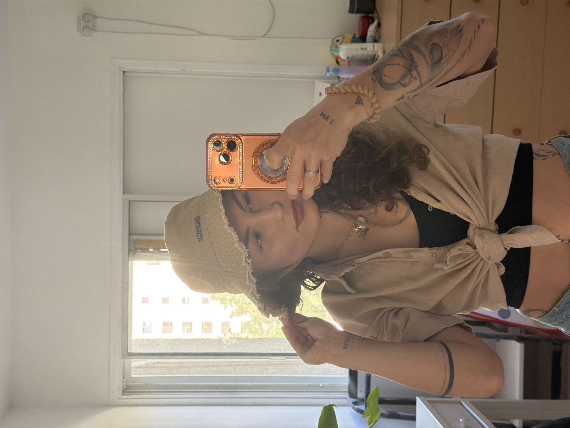 An indoor mirror selfie captures a young adult, likely female, with medium-length curly brown hair. She is wearing a light beige or tan button-up shirt tied at the waist over a black top, light blue jeans, and a matching light-colored bucket hat. She holds a rose gold smartphone with a jeweled case and a pop socket in her left hand, taking the photograph. Her right hand is near her ear, subtly touching her hair.
The subject displays several tattoos: on her left forearm, the text "MA I" (or similar) and a small triangle symbol are visible. Her left upper arm has a larger, intricate design, possibly an eye or abstract pattern. On her right forearm, the text "DIYAANS" (or similar) is tattooed, and a solid black band is on her right upper arm. A tattoo resembling a feather or wing is on her right rib/torso area. She is wearing a ring on her left hand, a light-colored beaded bracelet, and a necklace with a charm.
The background reveals a white room with a window on the left. Bright daylight streams through the window, illuminating a multi-story building outside, suggesting an urban or residential setting. An electrical outlet with wires is visible on the upper left wall. Some household items are vaguely discernible on a shelf in the upper right. A green plant leaf is partially visible at the bottom left. The text "TREYMOORE" is visible, inverted due to the mirror reflection, on the front of her bucket hat. The scene suggests a sunny daytime in Kiryat Bialik, Israel.