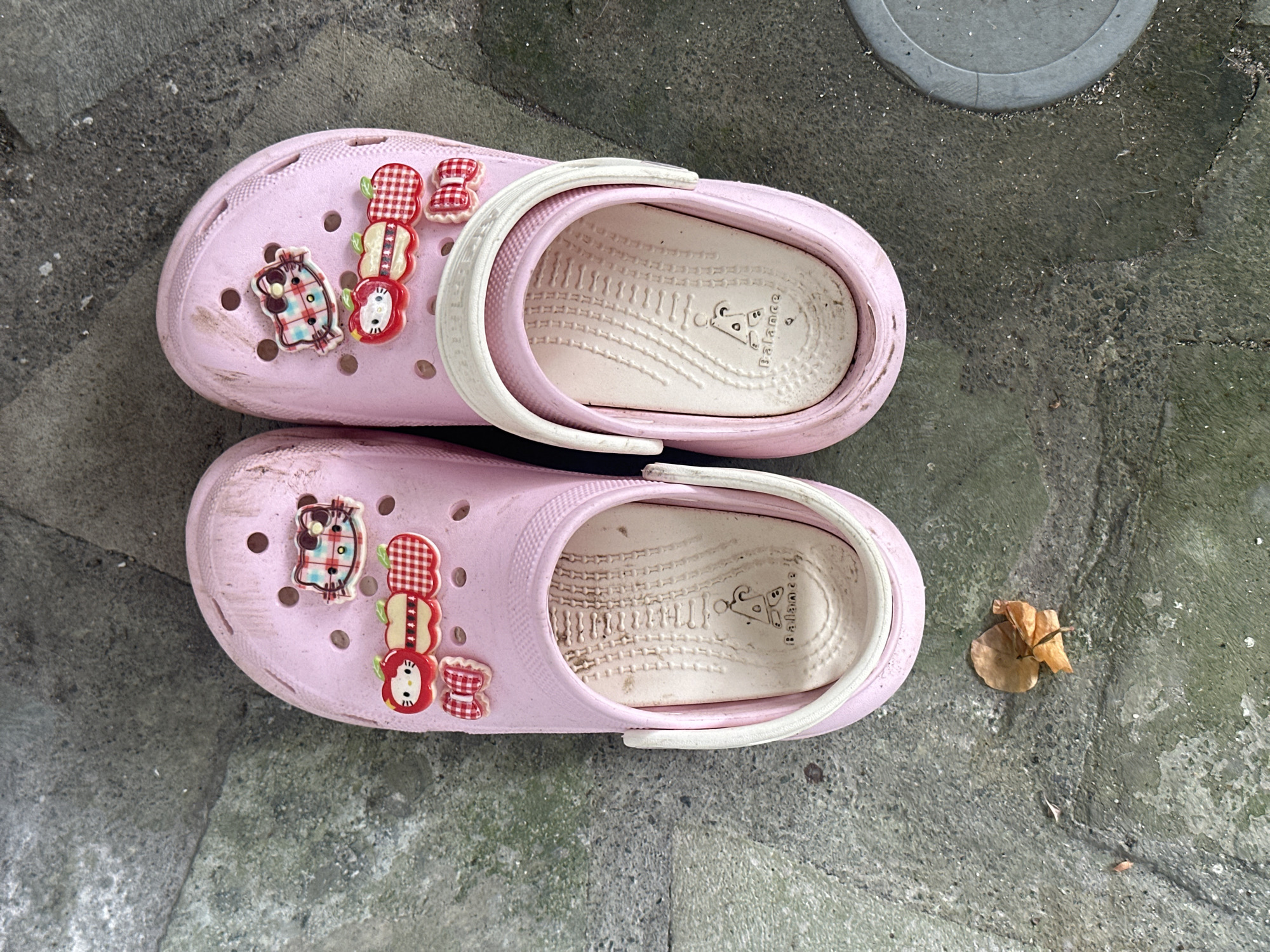 The image depicts a pair of pink Croc-style sandals adorned with cartoon character embellishments and red and white gingham bows. The footwear appears worn and dusty, resting on a weathered grey concrete surface. A dried leaf lies near the sandals. The setting appears to be outdoors. The city is North Kuta, Indonesia.