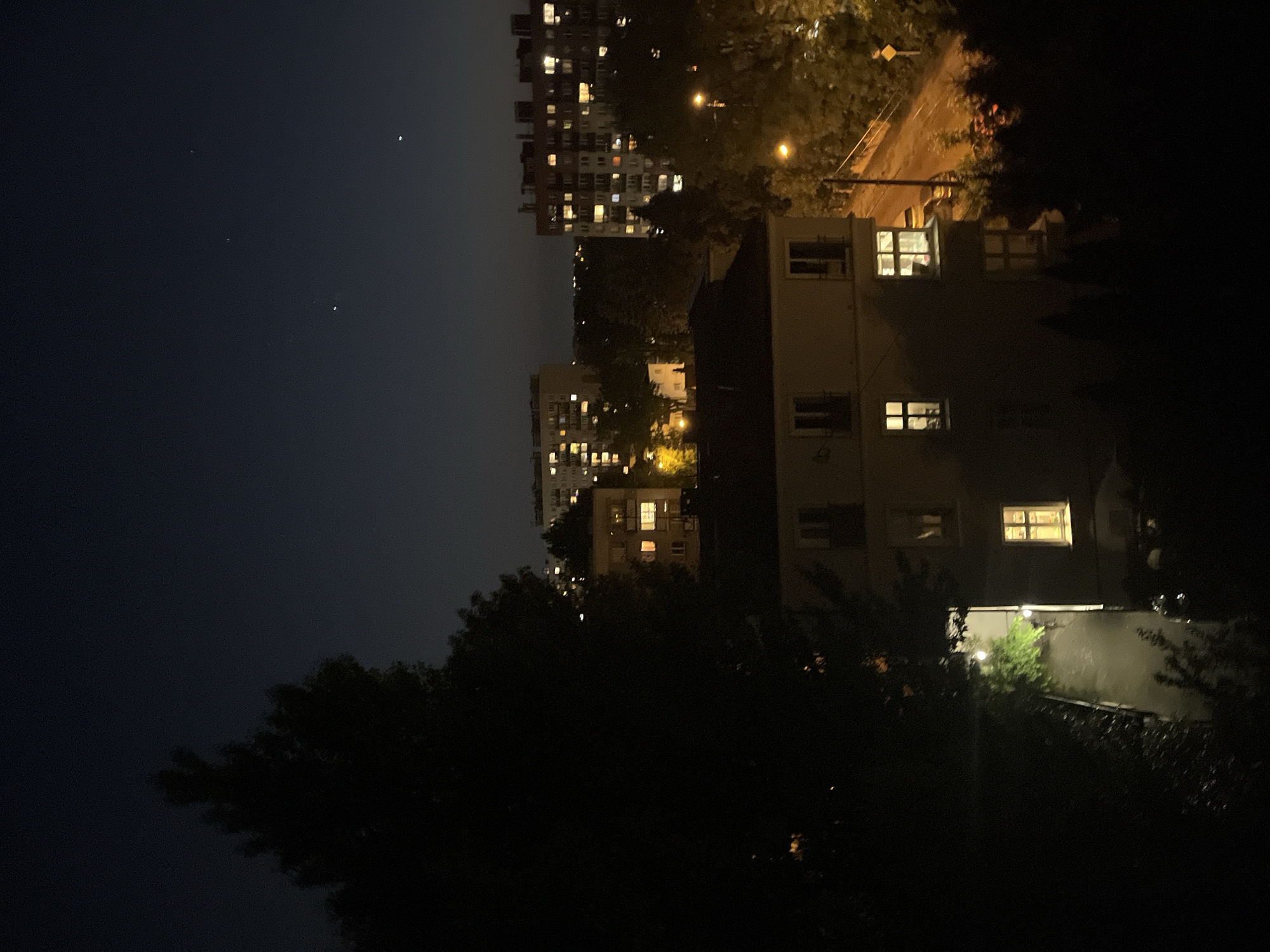 The image shows an urban nighttime scene with several buildings. One large apartment building with many lit windows is visible in the background. A smaller building is closer to the viewer, also with several lit windows. Trees are present in the foreground, appearing dark due to the lack of light. The sky is dark, with a few small points of light possibly indicating stars. A street lamp or other light source is visible in the middle ground, illuminating a path or road.