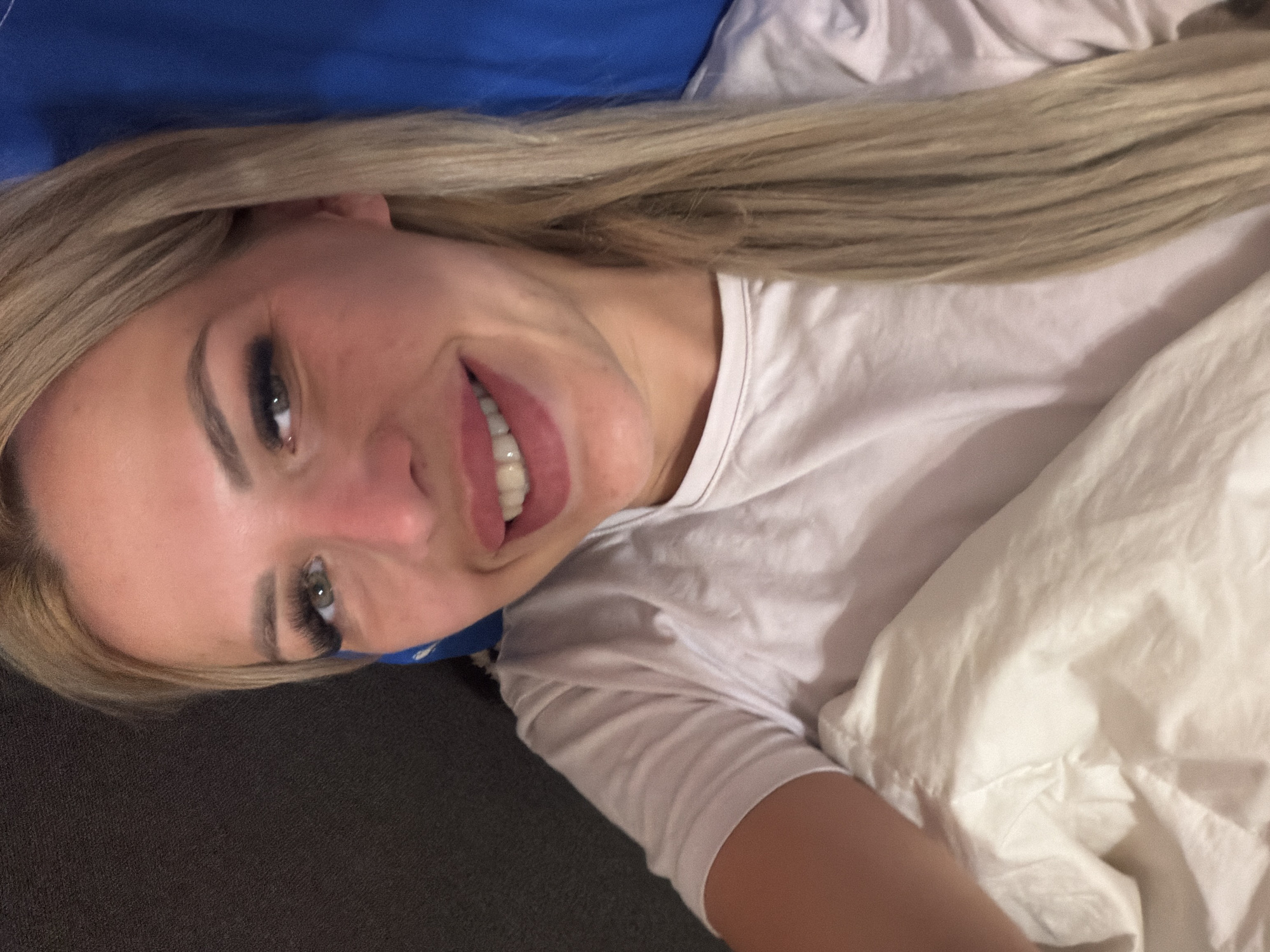 The image shows a close-up, sideways view of a young woman lying in bed. She has long, blonde hair and is wearing a white t-shirt. Her eyes are open, and she has a slight smile on her face. The background consists of a blue pillow and white bedding. The overall impression is one of relaxation and comfort, possibly in the morning or evening. There is no visible text in the image.