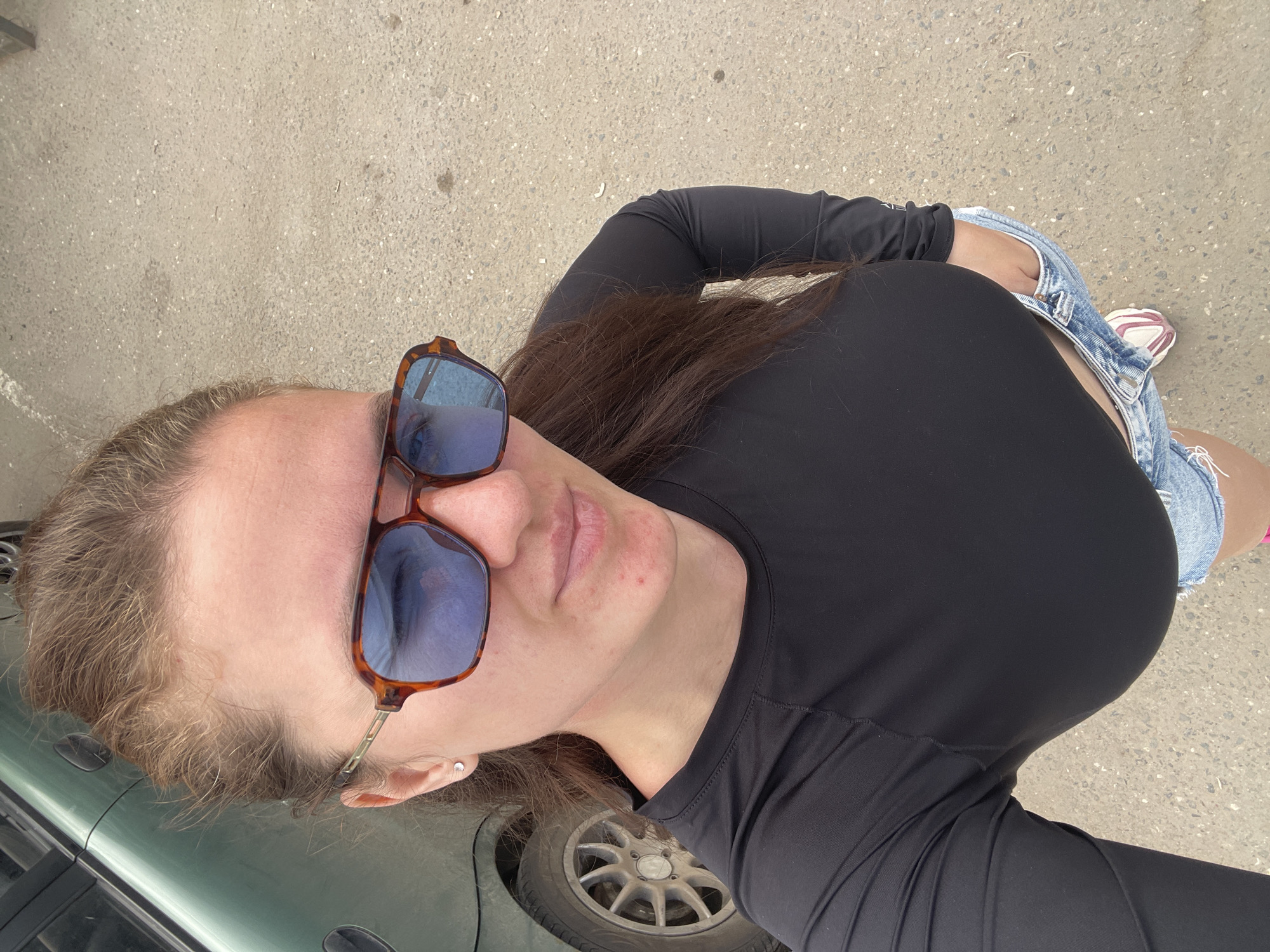 A woman wearing sunglasses, a black long-sleeved shirt, and denim shorts stands on what appears to be an asphalt surface, possibly a road or parking area. A car, predominantly green, is visible near her. The setting might be the city of Molokovo, Russia, but there are no distinct local landmarks visible in the image to confirm this.