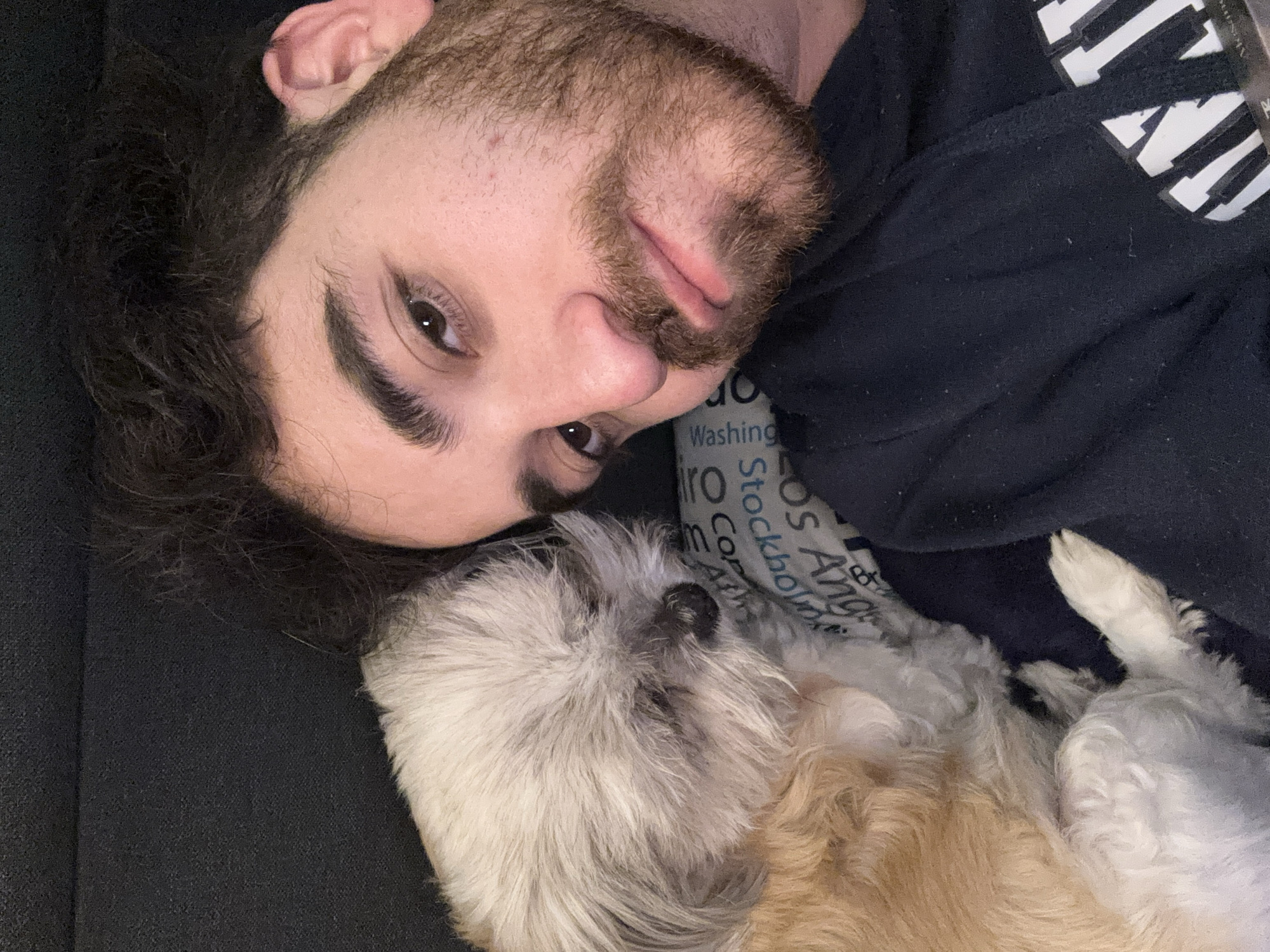 A man is lying down, with his head turned slightly to the left and resting near a small dog. The man has brown hair and a beard. The dog appears to be a Shih Tzu mix, with light brown and gray fur. They are both resting on a dark gray or black surface, possibly a couch or bed. Behind the dog is a pillow with text printed on it, but the details are unreadable. The man is wearing a dark-colored shirt, part of which is visible at the top right of the frame. The exact location is not discernable from this image and cannot be determined to be Santa Clara Ocoyucan, Mexico.
