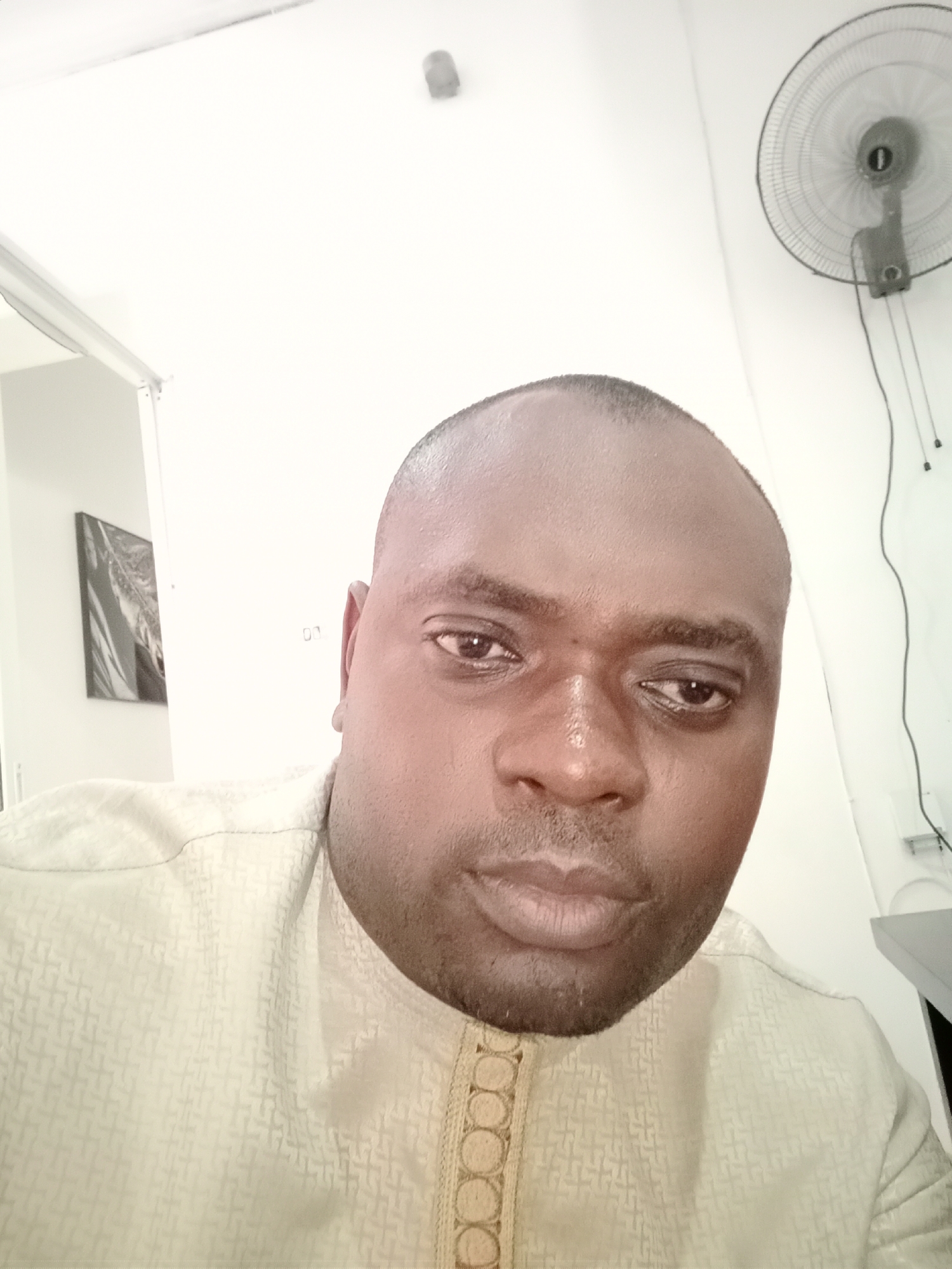 A close-up of a bald, dark-skinned man wearing a patterned beige shirt with an ornamental trim around the collar. Behind him is a white wall with an electrical outlet, a wall-mounted fan, and a doorway leading to a room with a framed picture. There is not enough information to confirm the location in the city of Gwarinpa, Nigeria.