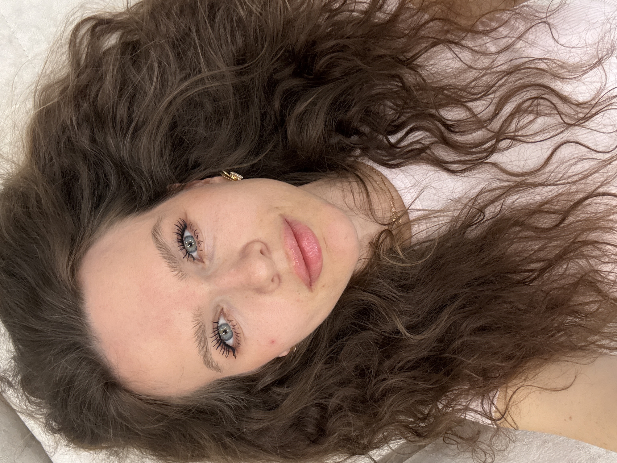 A woman with light skin, light blue eyes, and dark brown, wavy hair is lying down. Her head is surrounded by her hair, and she is wearing mascara. A gold earring is visible in her left ear. A gold necklace is around her neck. She appears to be lying on a white surface, possibly a bed or soft padding. There is no evidence to suggest the location is Khimki, Russia.