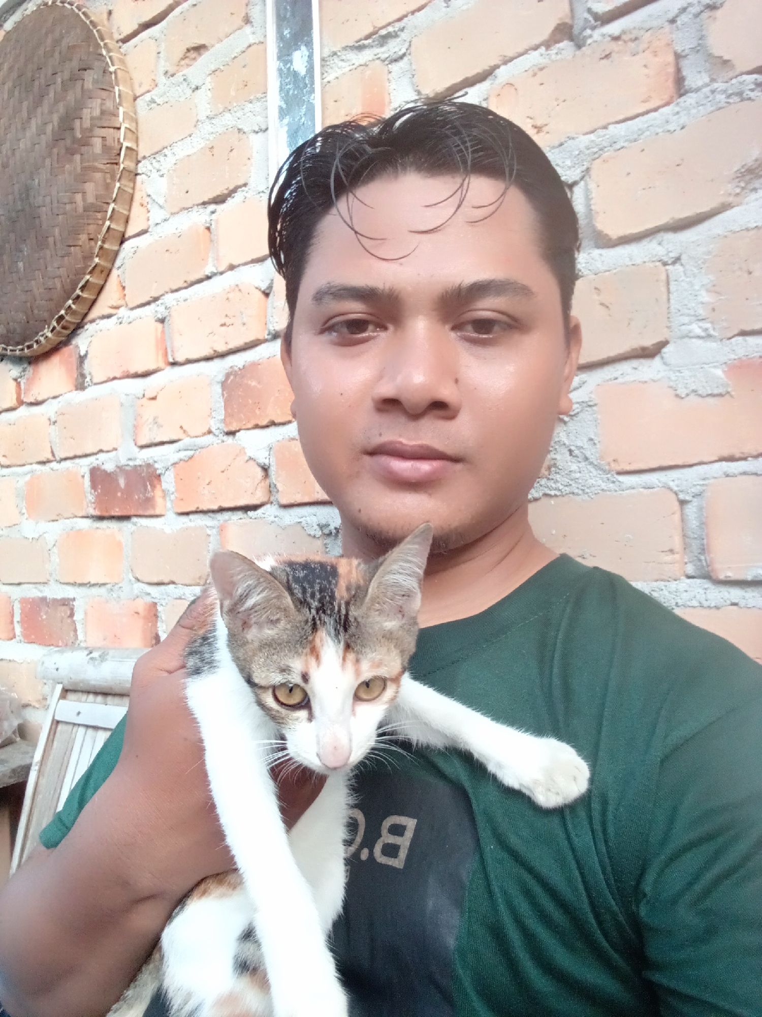 In Kota Negara Timur, Indonesia, a person with dark hair and medium skin tone is holding a calico cat in front of a brick wall. The person is wearing a dark green t-shirt. The cat, facing the camera, has white, brown, and black markings, with yellow eyes. To the left of the person, a woven bamboo circular object hangs on the brick wall. The environment appears to be outdoors, with part of a wooden chair visible at the bottom left. The lighting suggests daylight.