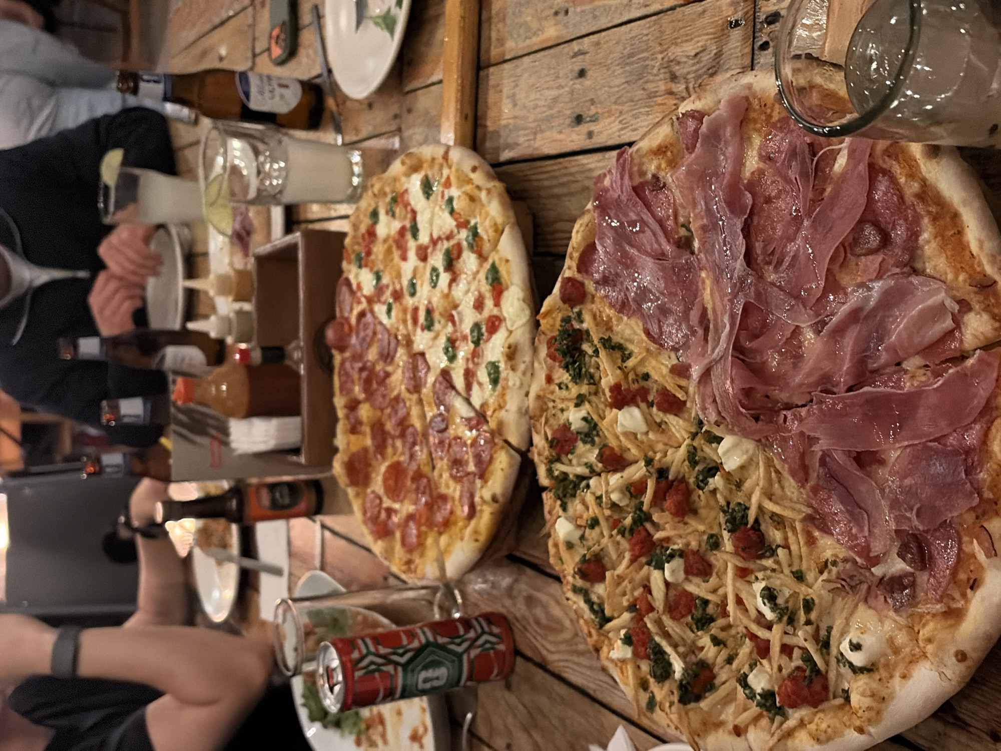 The image captures a lively social gathering, seemingly a meal, at an undetermined location in the city of Unknown, Unknown. A group of individuals surrounds a rustic wooden table laden with food and drinks. Two pizzas are prominently featured: one half pepperoni and half with basil and tomato, the other topped with what appears to be prosciutto and a mixture of smaller ingredients including green vegetable pieces, fried potato pieces, and white cream cheese. Clear glasses filled with beverages sit near the pizzas, along with beer bottles, various sauces and condiments, and a colorful can. Plates with leftover food are scattered around, suggesting an ongoing or recently concluded meal. People are visible in the periphery, engaging in conversation and eating. The lighting is warm, creating an inviting and convivial atmosphere.