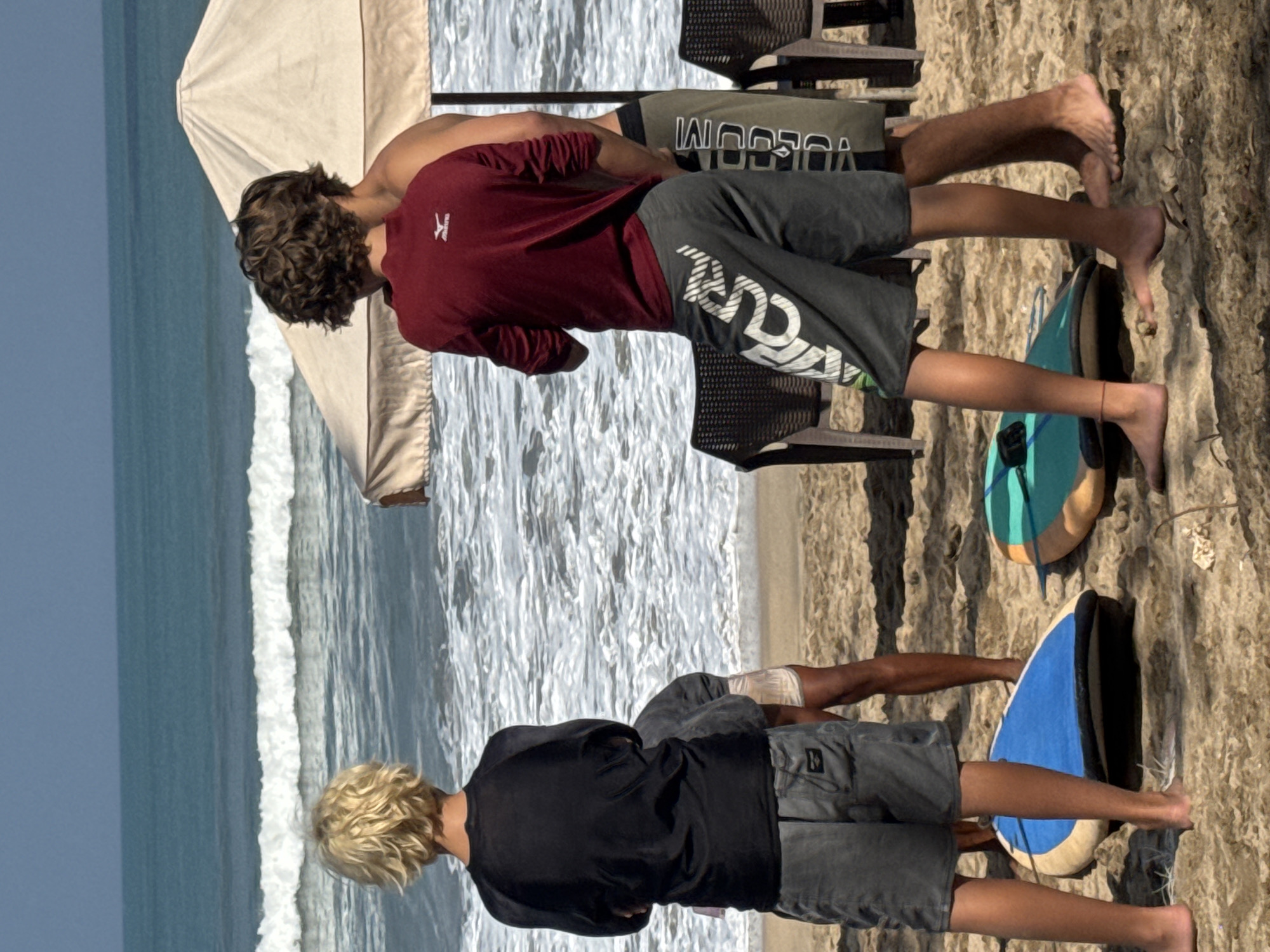 The image depicts a scene on a sandy beach, likely in Kuta, Indonesia, given the context. Three young males are visible, facing away from the camera towards the ocean. The ocean has moderate waves. In the foreground, two surfboards are lying on the sand, one blue and white, the other turquoise and orange. A tan beach umbrella is situated behind the two individuals in the center. Chairs with dark wicker backs are visible to the right.
The person on the left, with blond hair, wears a black long-sleeved rash guard and grey shorts. The person in the middle, with dark curly hair, wears a maroon long-sleeved shirt and grey shorts with white lettering. The person partially visible on the right wears dark shorts and appears to be standing next to the middle person. Their legs are visible, and they are barefoot. The sand is textured and shows footprints. The sky is clear and blue.