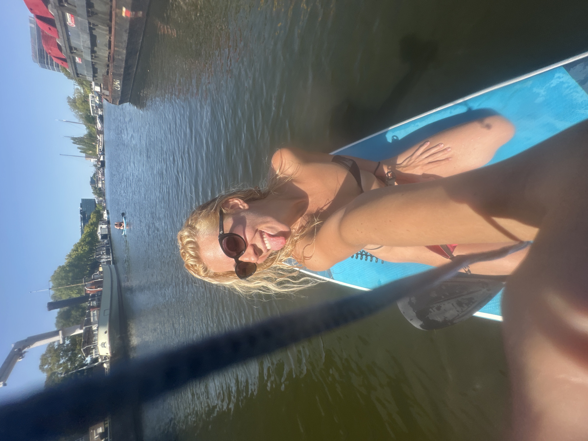 The image shows a woman on a paddleboard in a canal in Amsterdam, Netherlands. She is wearing sunglasses and a bikini, and her tongue is sticking out. The paddleboard is blue and white. In the background, there are several boats and buildings. The sky is clear and blue. The canal appears to be busy with other boats.