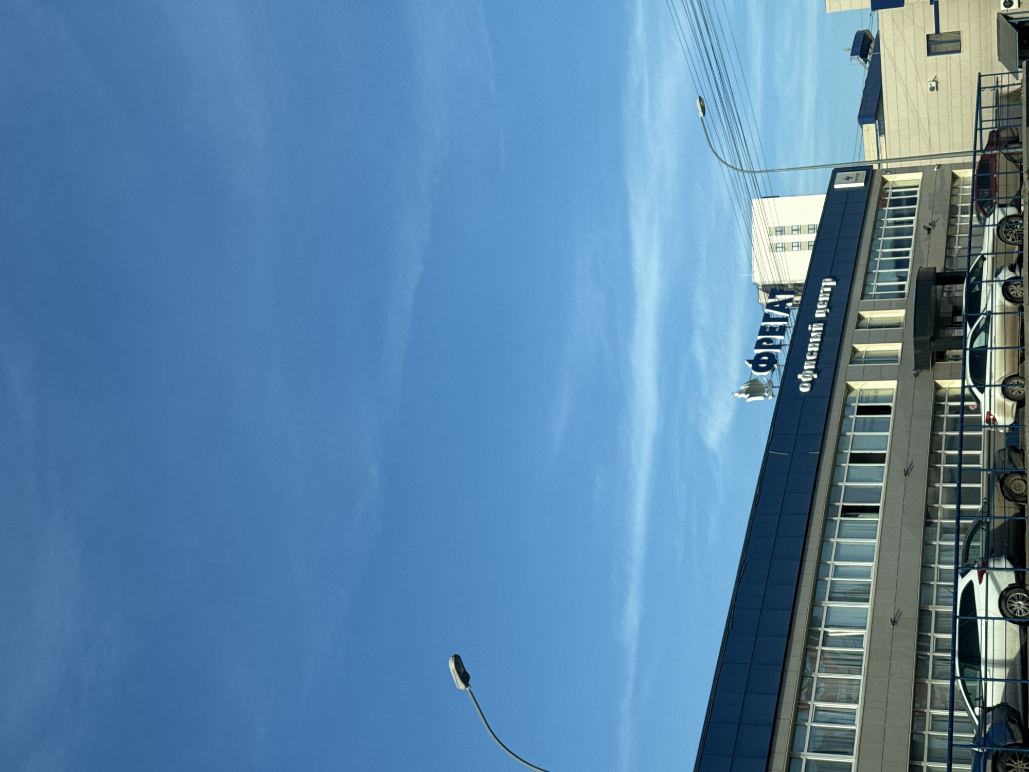 The image displays an exterior view of the "FREGAT" office center, located at Ulitsa Pushkinskaya, 240, Izhevsk, Russia. The scene is illuminated by daylight under a clear blue sky with minimal, wispy clouds. The multi-story building features a dark blue paneling on its upper section and roof, with the lower facade constructed of light gray or beige materials and lined with numerous windows. Several second-story windows are partially open. A large sign in blue Cyrillic letters identifies the building as "ФРЕГАТ" (FREGAT), with "офисный центр" (office center) displayed below it. A stylized white bird or sailboat figure surmounts the "ФРЕГАТ" lettering. A small blue square emblem with a white cross is visible on the building's right-hand facade. In the foreground, a row of vehicles is parked behind a metal barrier, including a dark blue car (partial view), a white station wagon, a dark gray sedan, a maroon sedan, and another white car (partial view). No individuals are present. Streetlights with arcing poles are visible on both the left and right sides of the frame, with utility lines crossing the upper right quadrant. In the background, a taller, light-colored building with multiple windows and a beige, textured building with rooftop equipment are discernible.