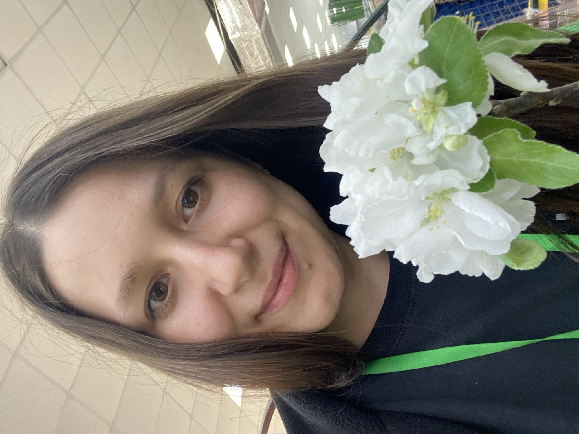 A woman with long, dark brown hair is holding a cluster of white apple blossoms with green leaves. She is wearing a black shirt with a green strap. The background appears to be an interior space with a white tiled ceiling. While the exact location cannot be determined, the mention of Chaikovsky, Russia implies the photo was taken there, but it is not directly visible in the image.