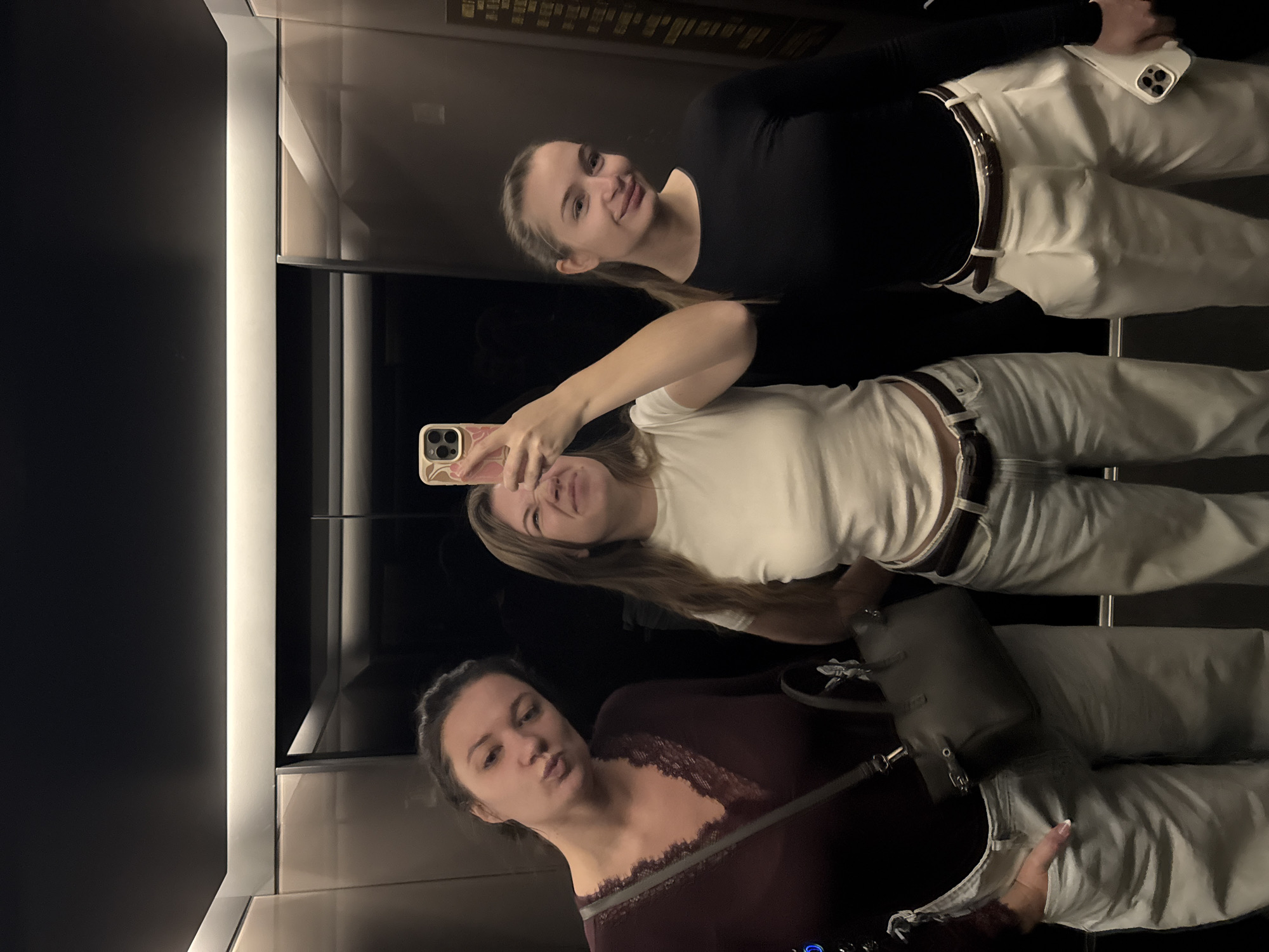 The image shows three women reflected in a mirror, likely in an elevator. The woman closest to the camera is wearing a dark red top with lace detailing. The middle woman is holding a phone, presumably taking the photo, and is wearing a white t-shirt. The third woman is wearing a black top. All three appear to be wearing white or light-colored pants with belts. A purse is visible beside the second woman. The elevator has dark panels. The precise location within the Vracar Urban Municipality, Serbia is not discernible from the image.