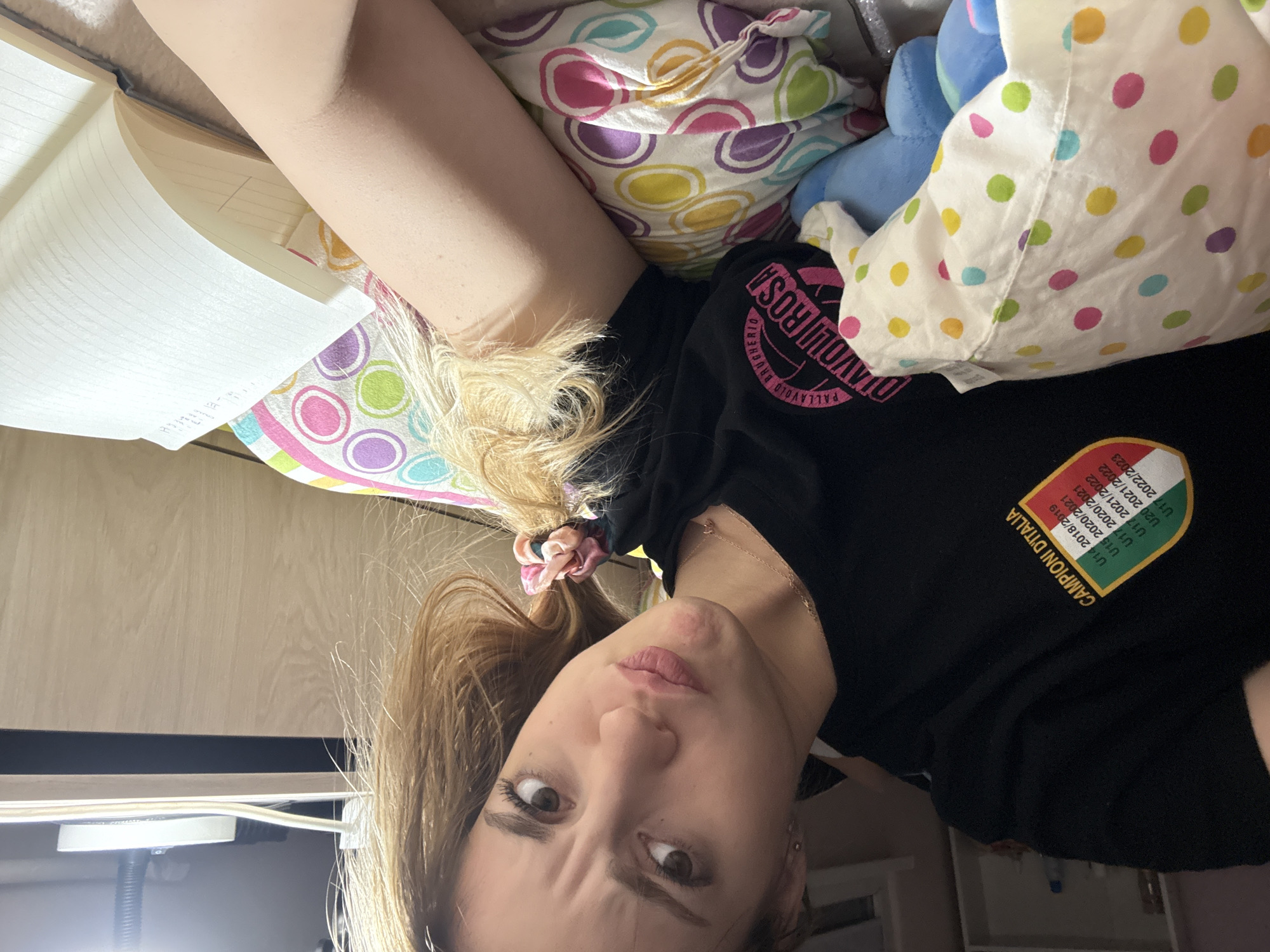 A woman is lying down, looking at the camera with a slightly quizzical expression. She has fair skin, brown eyes, and blonde/brown hair pulled back with a scrunchie. She is wearing a black t-shirt with the words "DIAVOLI ROSA PALLAVOLO BRUGHERIO" and a patch that reads "CAMPIONI D'ITALIA" along with various years. Her head rests on a pillow with multicolored circles, and there is another pillow with polka dots behind her. A book is visible to the left, along with a desk lamp. The scene appears to be indoors. There is no indication that this image was taken in Sofia, Bulgaria.