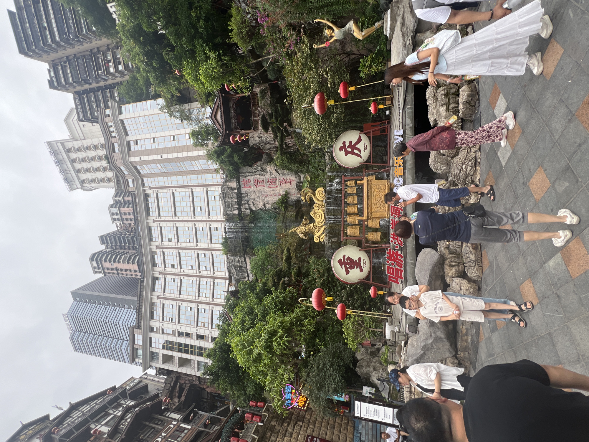The image shows a street scene in Yuzhong District, China, featuring a multi-story building complex with traditional Chinese architectural elements and modern high-rise buildings in the background. In the foreground, several people are standing and walking on a paved walkway. A central feature is a display with large red drums, musical instruments, and decorative golden dragon sculptures, situated against a rocky, landscaped backdrop with a waterfall. Red lanterns are hung throughout the scene. A statue of a woman in a dancing pose is visible on the right side. The sky is overcast.