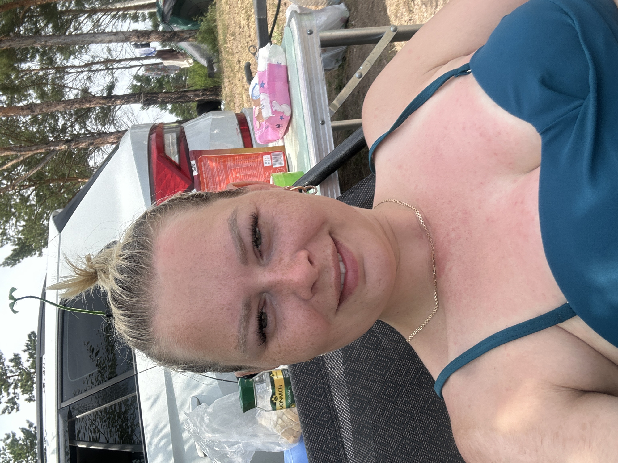 A woman with blonde hair tied in a bun, wearing a teal bikini top and a gold necklace, is photographed in a close-up selfie. She is outdoors, with a white SUV visible behind her, and a picnic table in the background laden with items including a pack of diapers and a jar of Monarch Original spices. The environment appears to be a campsite, with trees and possibly tents in the distance, suggesting a rural or forested setting. The woman's skin shows signs of sun exposure with redness on her chest. A thin green stem with two leaves is stuck in her hair. The image was captured in Kolodeznyy, Russia.