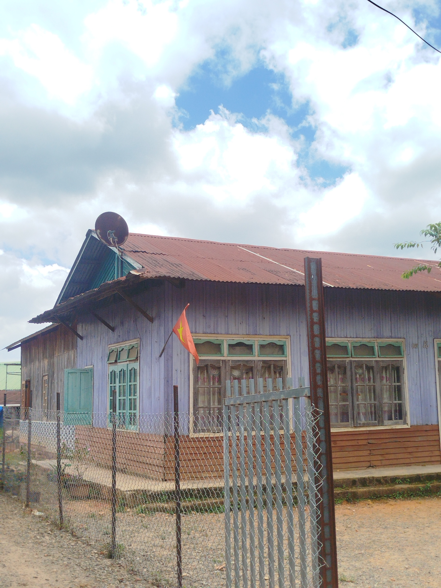 The image shows a weathered, light purple wooden building with a rusty red corrugated metal roof, situated in Thôn 6, Vietnam. The building has several windows with light green frames and what appear to be older, possibly glassless panes. A Vietnamese flag is displayed on a pole extending from the building. A large, dark, dish-like object is mounted on the peak of the roof. A chain-link fence with metal posts runs across the foreground, obscuring part of the building's lower half. The ground is unpaved and covered with gravel and dirt. The sky is partly cloudy with patches of blue. The overall impression is one of a rural or semi-rural setting with a building that has seen better days.