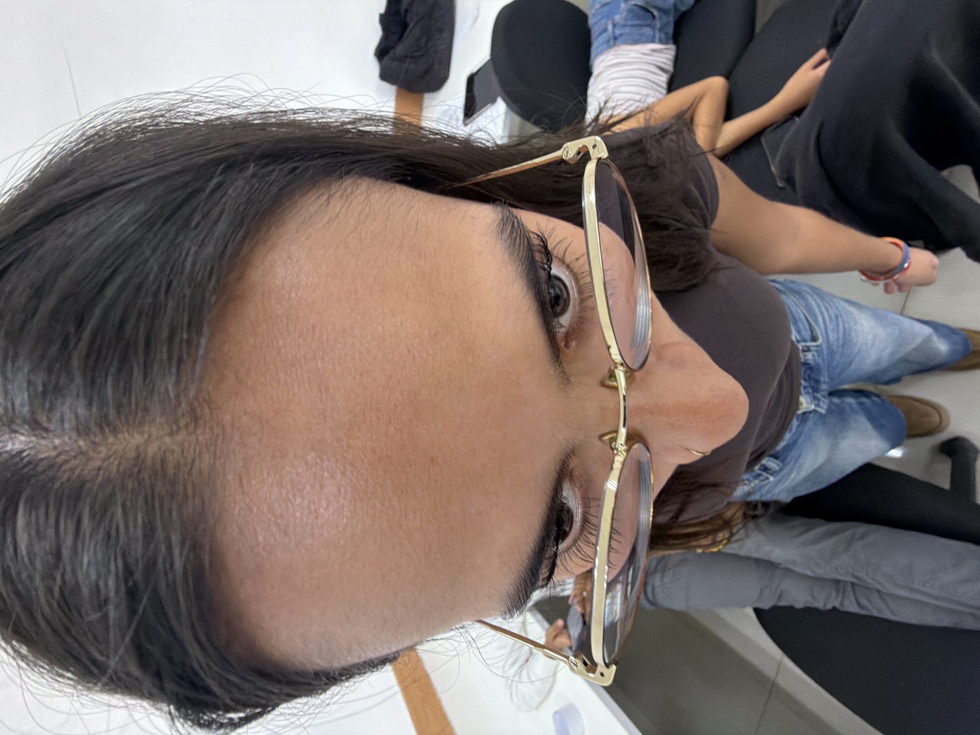 An overhead, close-up shot features a woman with dark hair and gold-rimmed, round glasses, looking upwards and slightly to her right. The primary focus is on her forehead, right eye with prominent eyelashes, and nose, while the rest of her face is out of frame. She is wearing a dark brown t-shirt and blue denim jeans.
In the softly blurred background, the limbs and torsos of at least two other individuals are visible, suggesting an indoor, casual gathering. One person, possibly a child, wears a striped t-shirt and has an arm resting, while another person is seen wearing black leggings. A colorful red and blue wristband is visible on one of the arms in the background. Other objects include what appears to be a dark phone or tablet on a surface, and dark shoes or boots.
The lighting is even, suggesting daytime. The setting appears to be an indoor space with white surfaces and dark furniture, implying a relaxed environment. There is no visible text in the image. The context for the photo is San Andrés Cholula, Mexico.