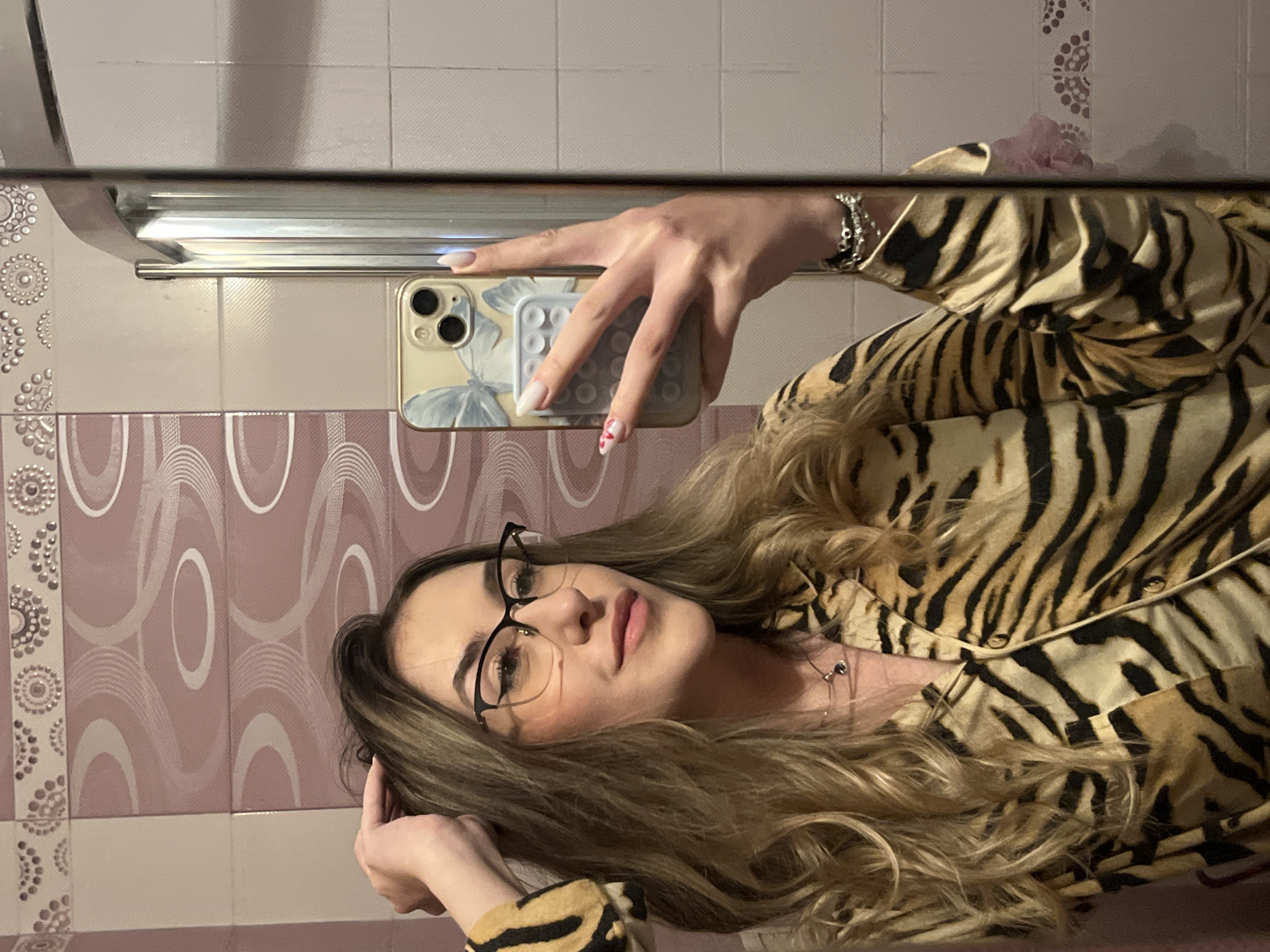 A woman is taking a selfie in a bathroom mirror. She is wearing glasses and a tiger-striped shirt. She is holding a smartphone with a butterfly-patterned case. The bathroom has mauve and white patterned tiles. It is not possible to determine the location within the city of Sofia, Bulgaria, or the precise interactions or actions taking place beyond the selfie.