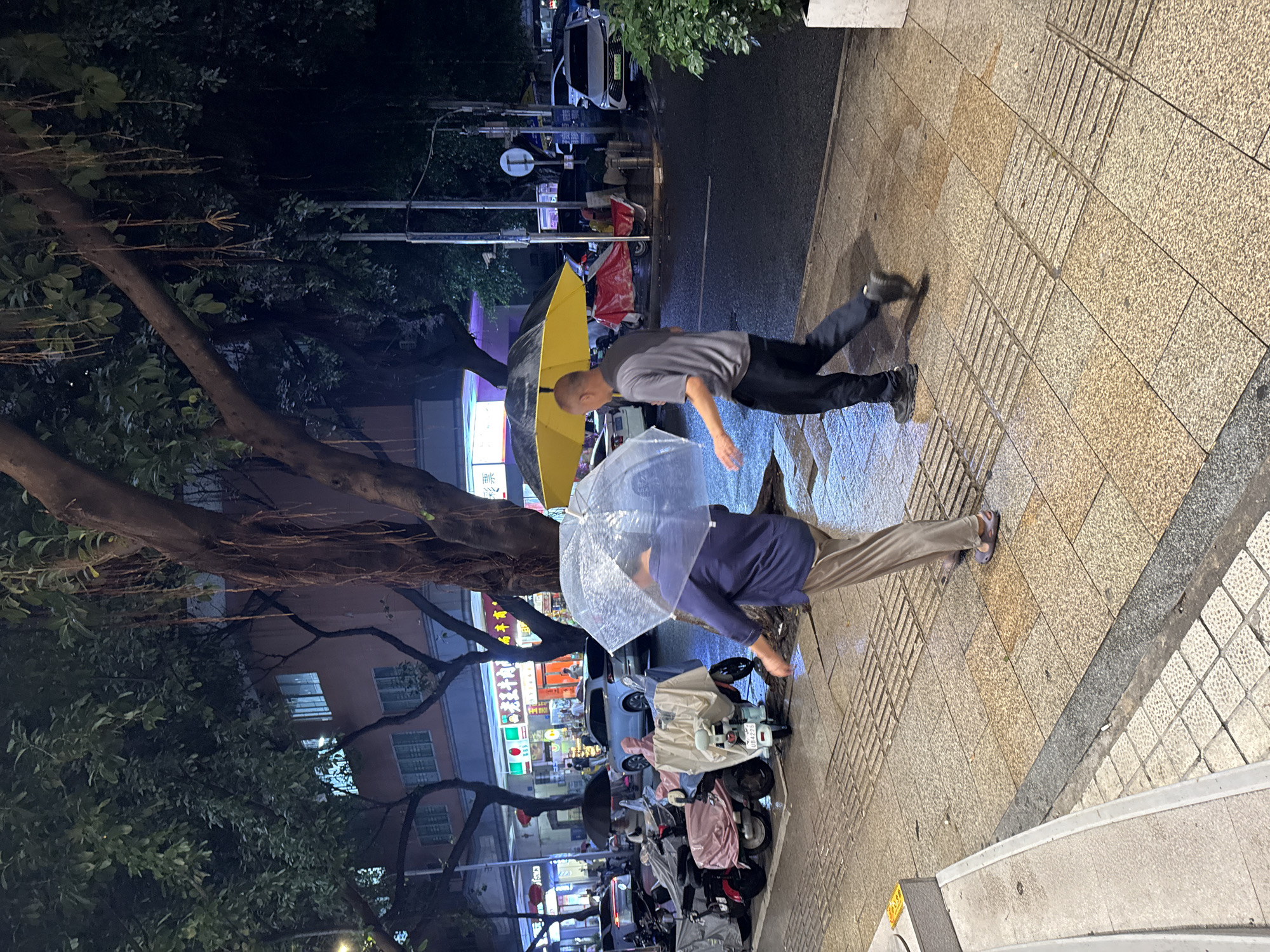 The image shows a street scene at night, likely in a city in the Tianhe District, China, given the presence of Chinese signage. The focus is on two people walking on the sidewalk. One person is carrying a yellow umbrella and is wearing a gray t-shirt and black pants. The other person has a clear umbrella and is wearing a blue long sleeve shirt and light colored pants. They are walking on a wet sidewalk, which suggests it has been raining. Along the side of the road are multiple parked motor scooters covered with various tarps and covers. There are trees on the left and buildings and shops in the background, with signs written in Chinese.