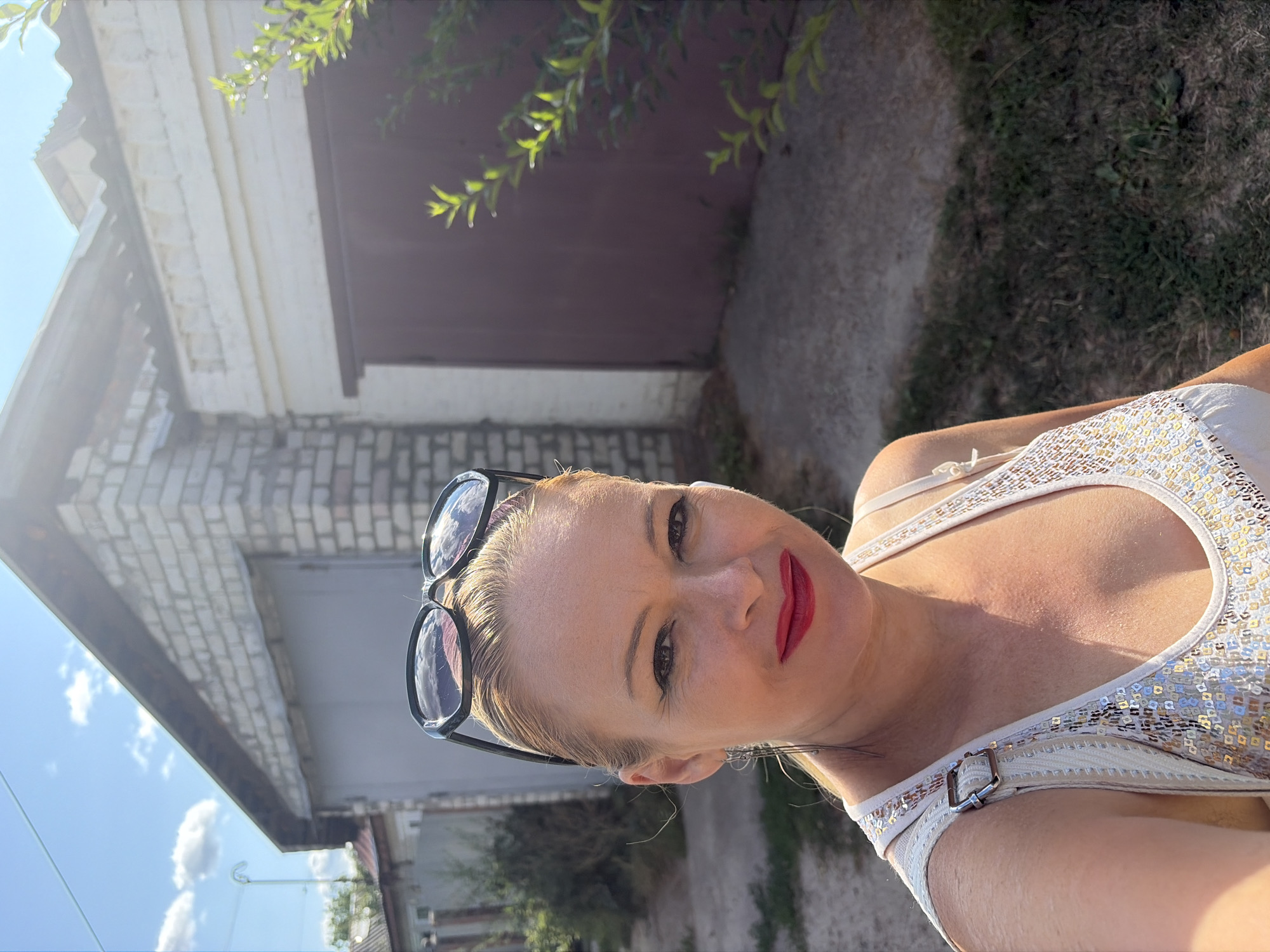 A woman with blonde hair, wearing sunglasses on her head, a white sequined tank top, and red lipstick, is taking a selfie outdoors. She has on earrings. In the background, there are garages, one made of brick and another with a dark brown door, set against a bright, sunny sky with some clouds. Vegetation is visible on the right side of the frame. The ground in front of the garages is covered with dry grass and dirt. The scene appears to be in a residential or suburban area.