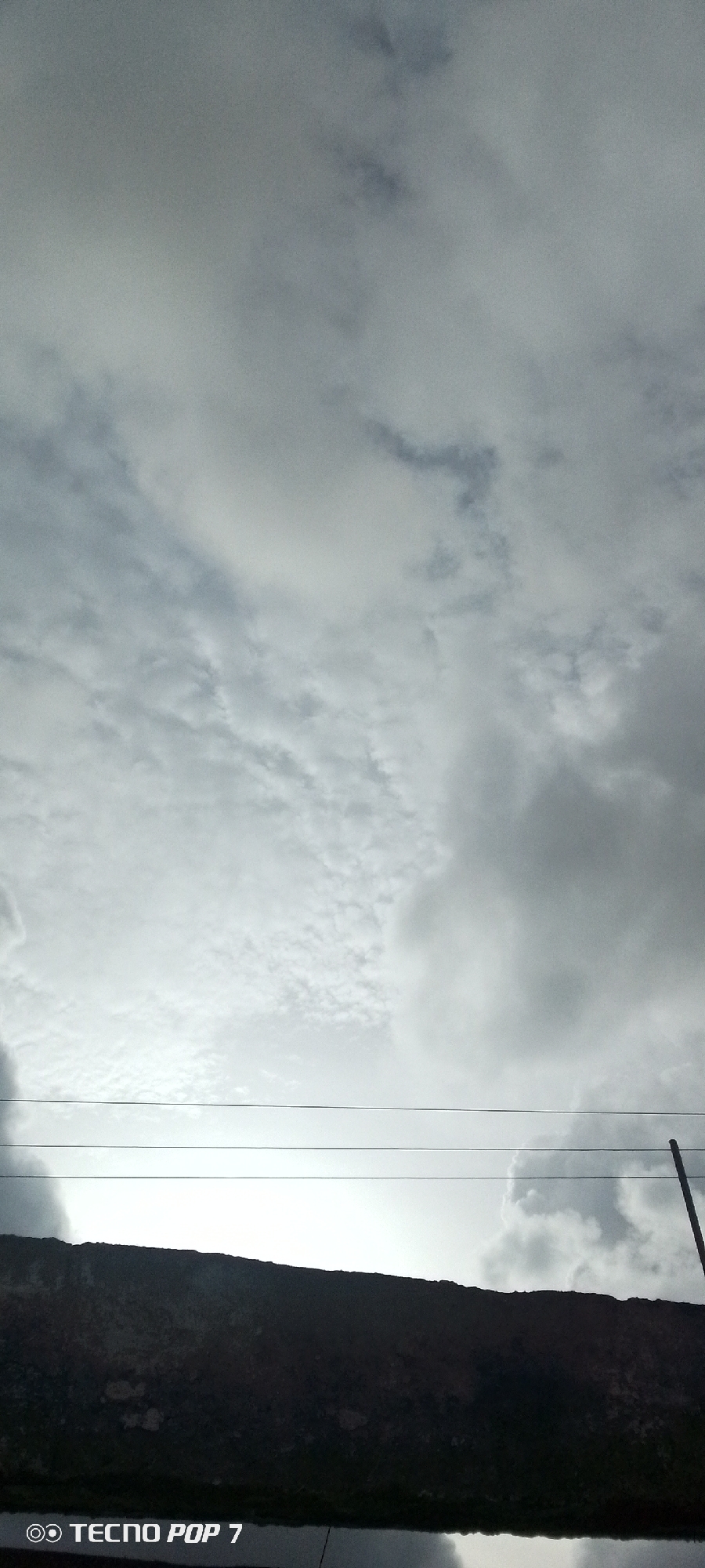 The image is a low-angle shot of a cloudy sky with a dark, textured structure in the foreground, possibly a wall or roofline. Three thin, dark lines, likely power or utility lines, stretch horizontally across the frame. A thin, vertical dark pole is visible on the right side of the image, partially obscured by the clouds. The bottom left corner of the image contains a watermark that reads "TECNO POP 7". The lighting suggests it is either early morning or late afternoon, or a heavily overcast day. The environment appears to be urban or semi-urban, as indicated by the utility lines and the constructed foreground element. The specific location in Altagracia, Venezuela, cannot be determined from the visual information alone, but the watermark suggests the image was captured using a Tecno Pop 7 smartphone.