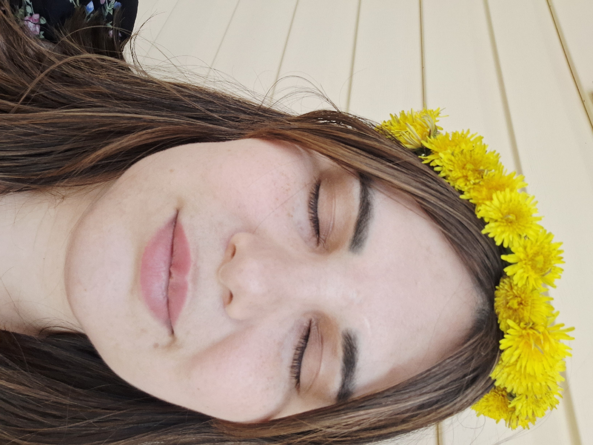 A close-up, horizontally oriented portrait depicts a woman with her eyes closed, wearing a crown of bright yellow flowers. Her hair is long, straight, and brown. Her skin appears fair and freckled. Her lips are closed and slightly parted. The background features a light-colored, vertically paneled surface. There is no indication from the image that this photo was taken in Амзя, Russia, or any other location.