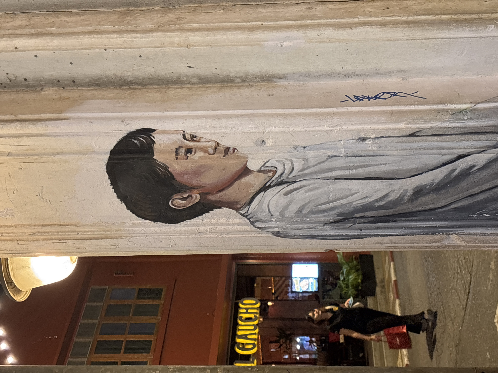 The image is of a mural of a person's face painted on a wall in Phuket City Municipality, Thailand. The person in the mural is lying down with their head turned to the right. The mural is on a light-colored wall with some texture. Above the mural is a signature. Below the mural, the image shows a portion of the street with a restaurant front visible. The restaurant sign reads "EL GAUCHO". A person is walking on the sidewalk. The lighting is dim.